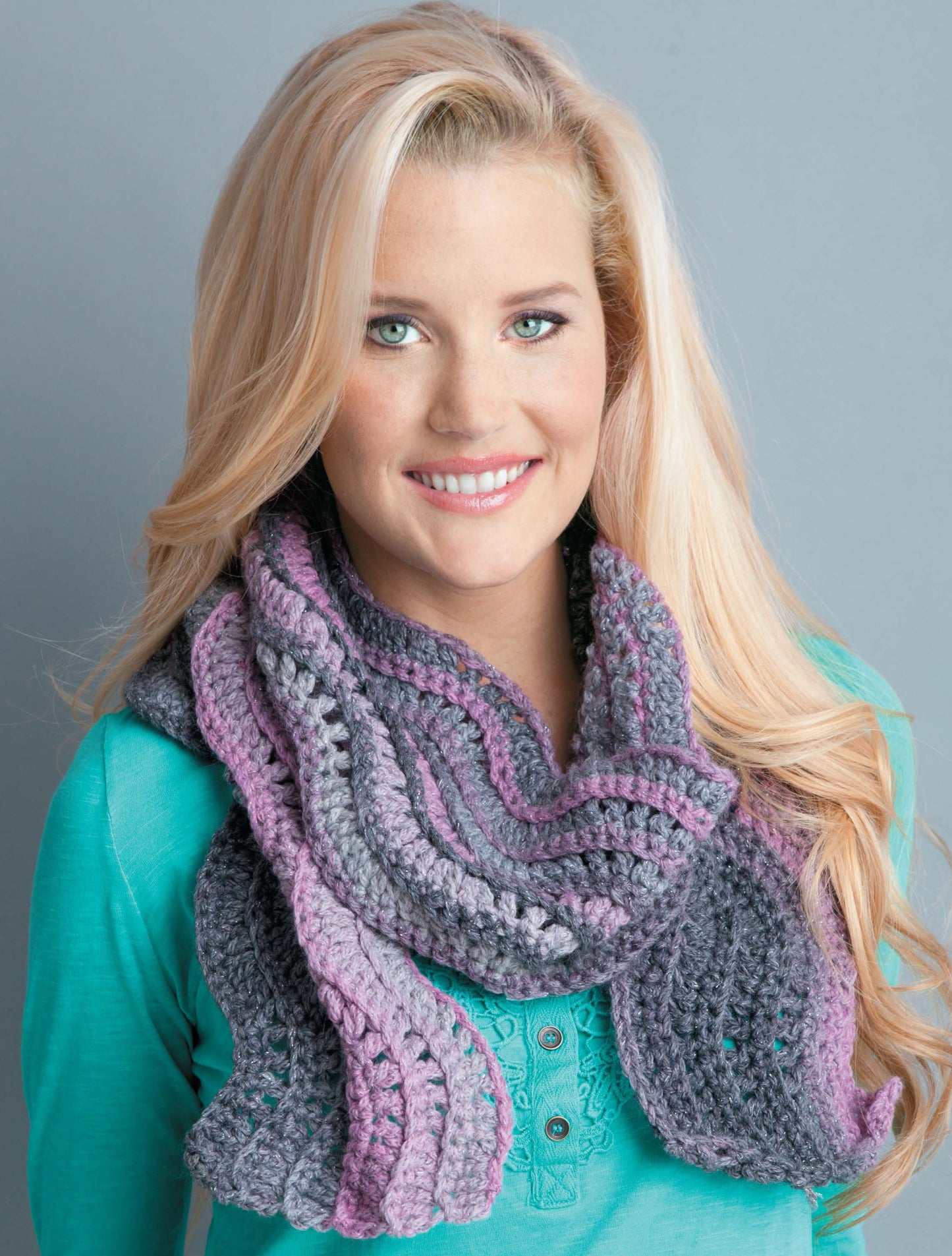 A woman with long blonde hair, wearing a teal top and a chunky purple, lilac, and gray scarf, smiles against a plain gray background—showcasing Leisure Arts’ "Crochet Scarves & Cowls" digital download for fresh crochet inspiration.
