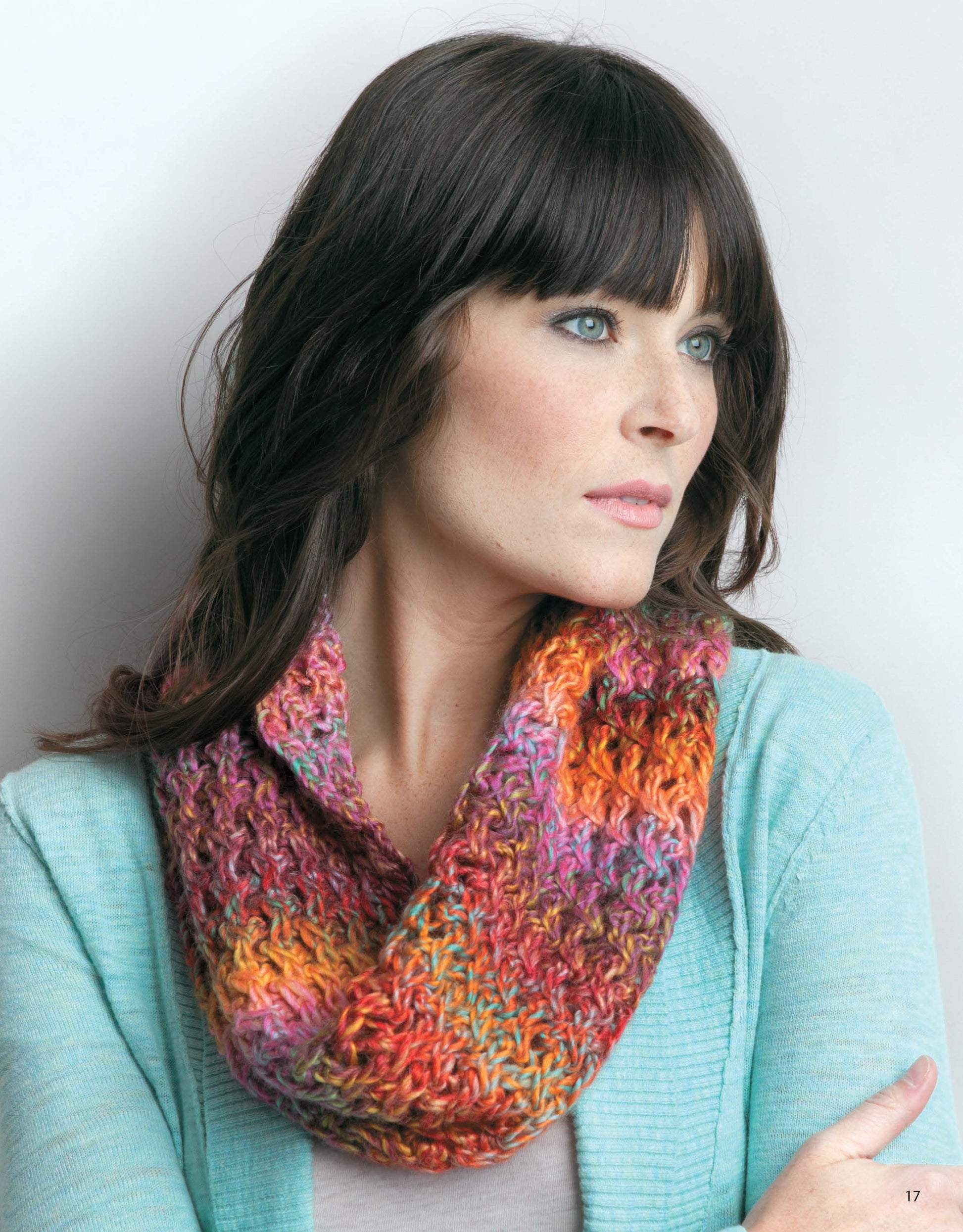 A woman with medium-length dark hair and bangs models a vibrant crocheted cowl from Knit Scarves & Cowls by Leisure Arts, layered over a light blue cardigan, as she gazes thoughtfully to the side against a light background.