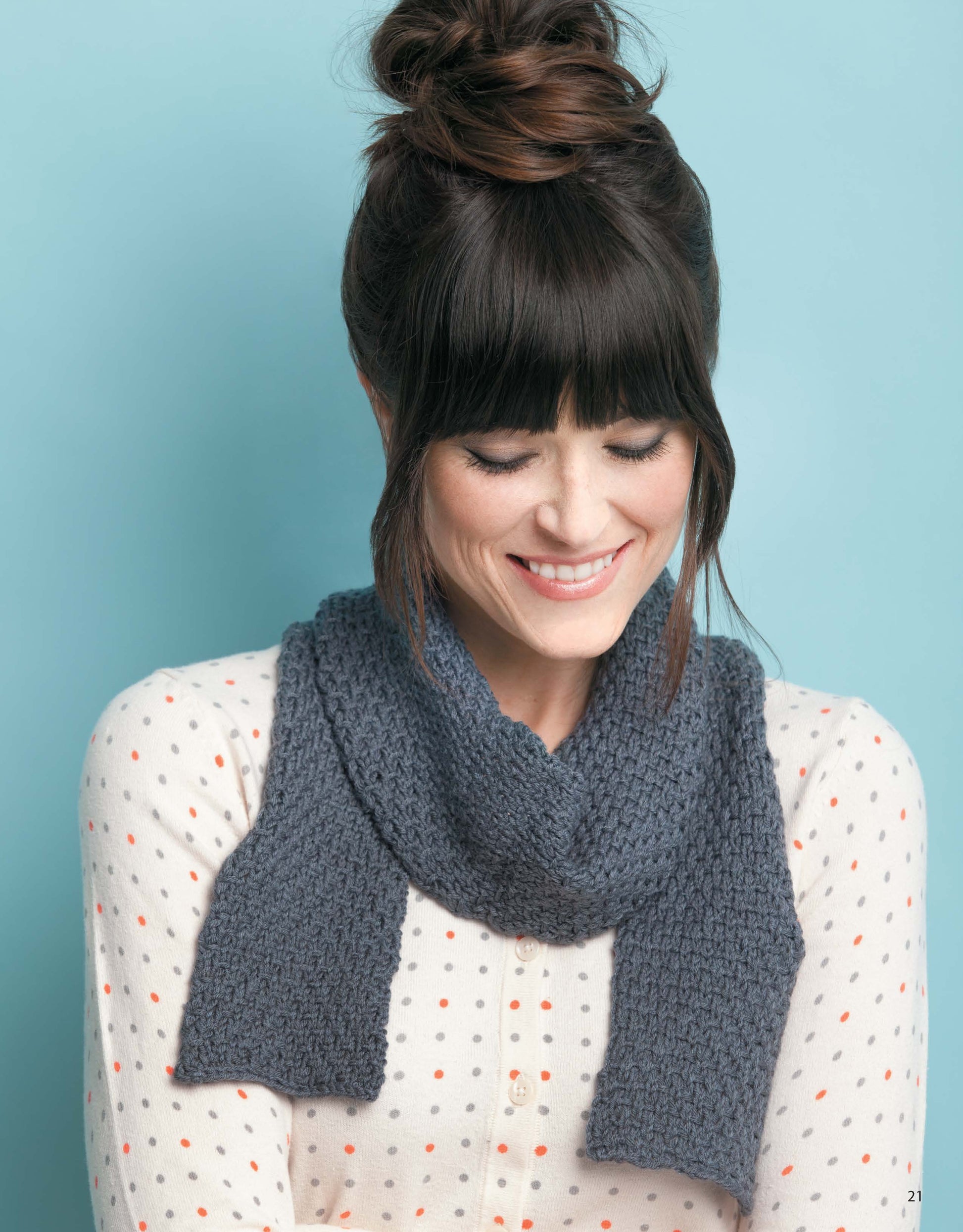 A woman with brown hair in a messy bun smiles, wearing a white shirt with orange and gray polka dots and a chunky gray cowl—great inspiration from Leisure Arts’ Knit Scarves & Cowls collection for anyone interested in stylish knitting patterns.