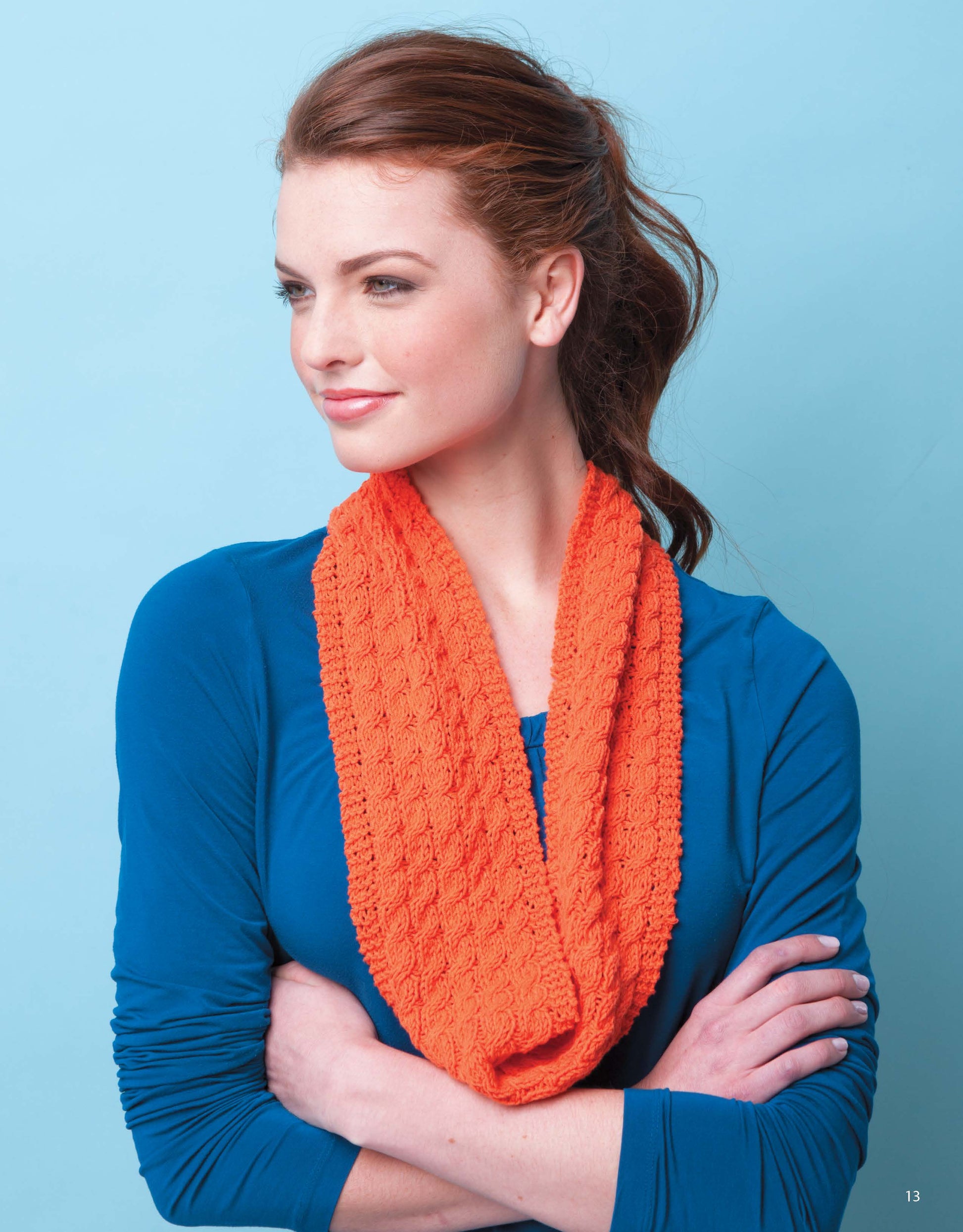 A woman with brown hair in a ponytail models a vibrant orange scarf from Leisure Arts' Knit Scarves & Cowls, paired with a blue long-sleeve top, standing arms crossed against a light blue background—a perfect beginner knitting inspiration.