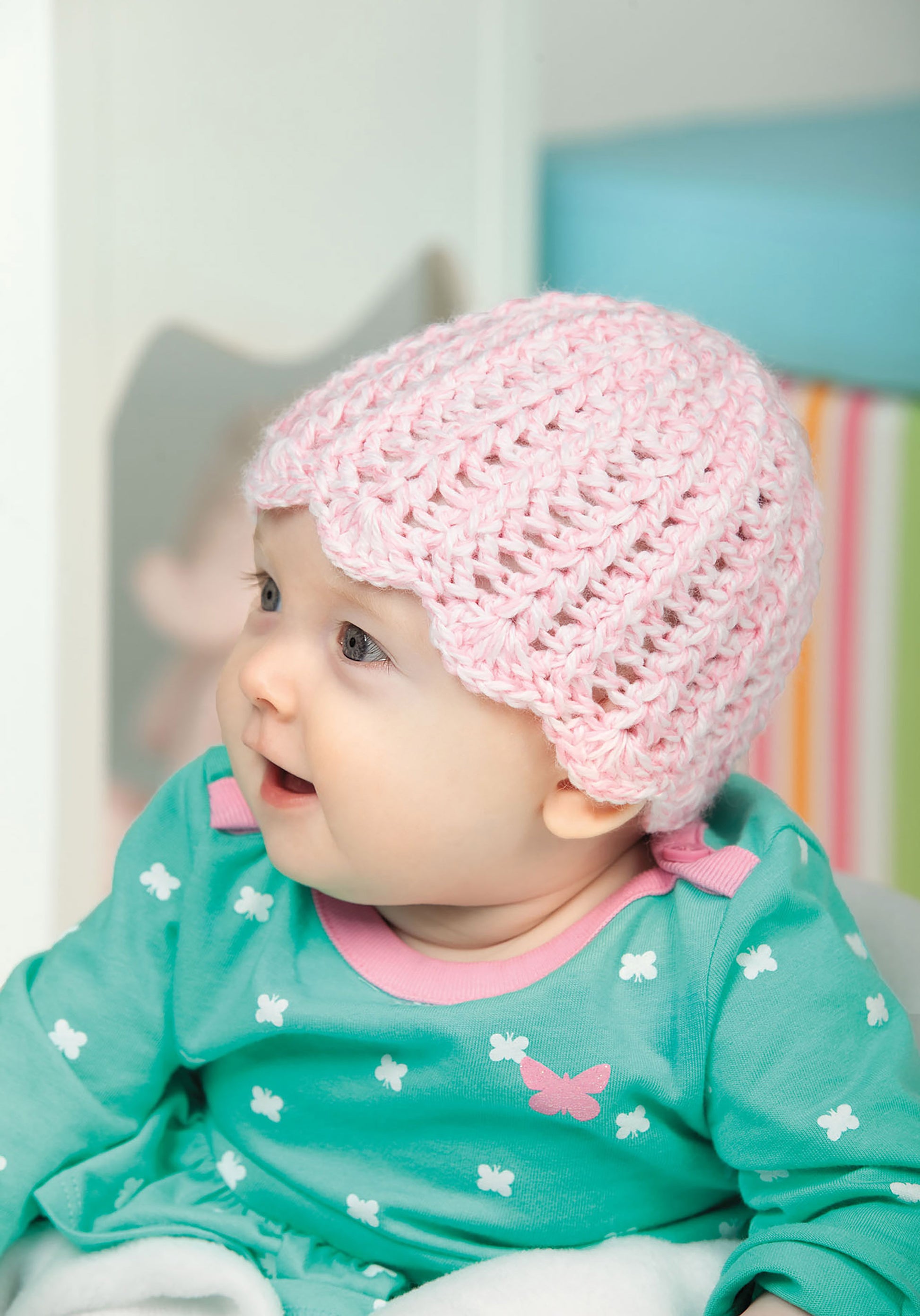 A baby wears a pink hat made from Leisure Arts’ “Baby Hats—10 Quick and Easy Patterns Written in Three Sizes” and a teal outfit with white bows and a pink butterfly, sitting indoors with a curious expression.