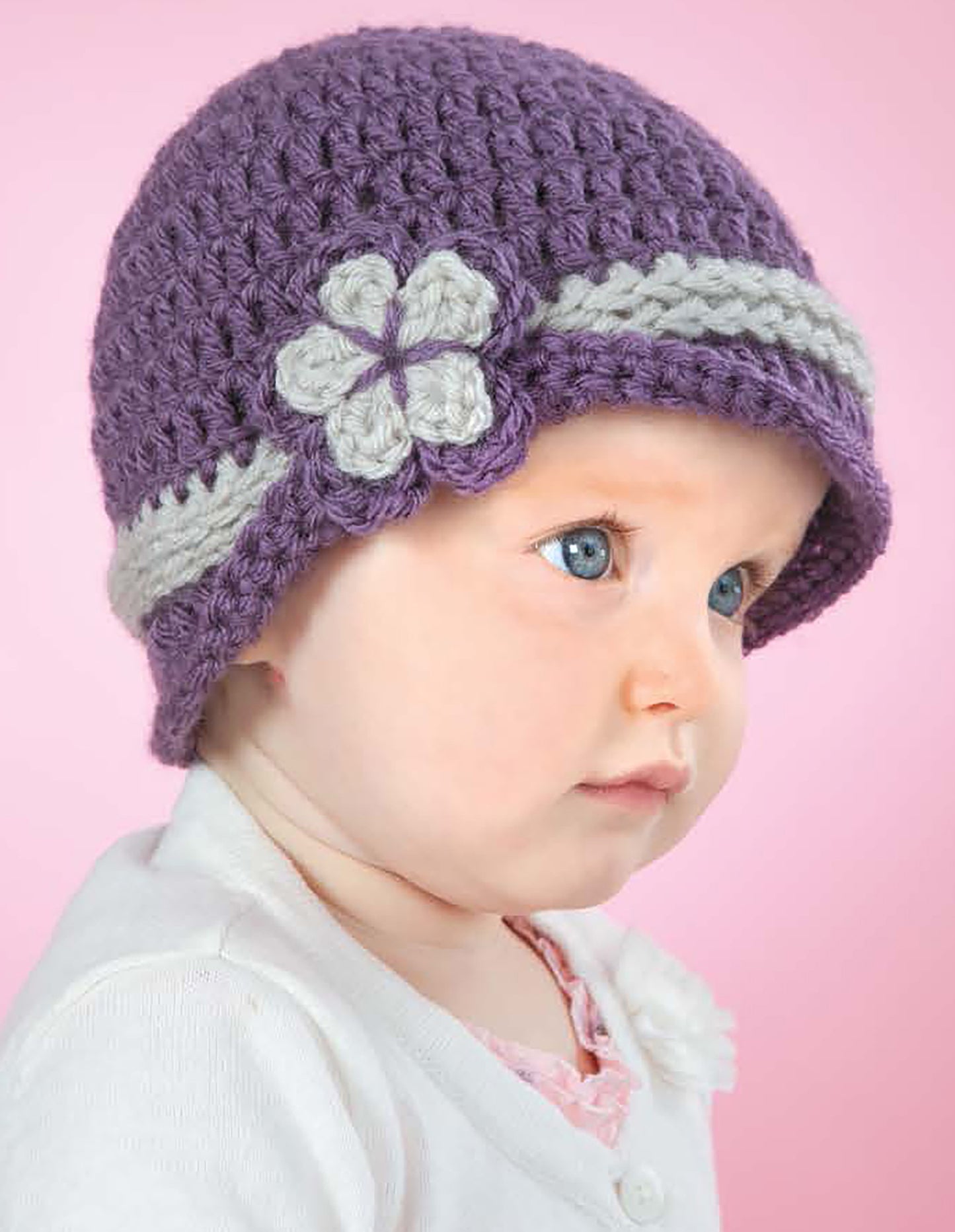 A baby in a white outfit wears a purple crocheted hat with a light gray band and flower, against a pink background. The hat is made using patterns from "Baby Hats—10 Quick and Easy Patterns Written in Three Sizes" by Leisure Arts.