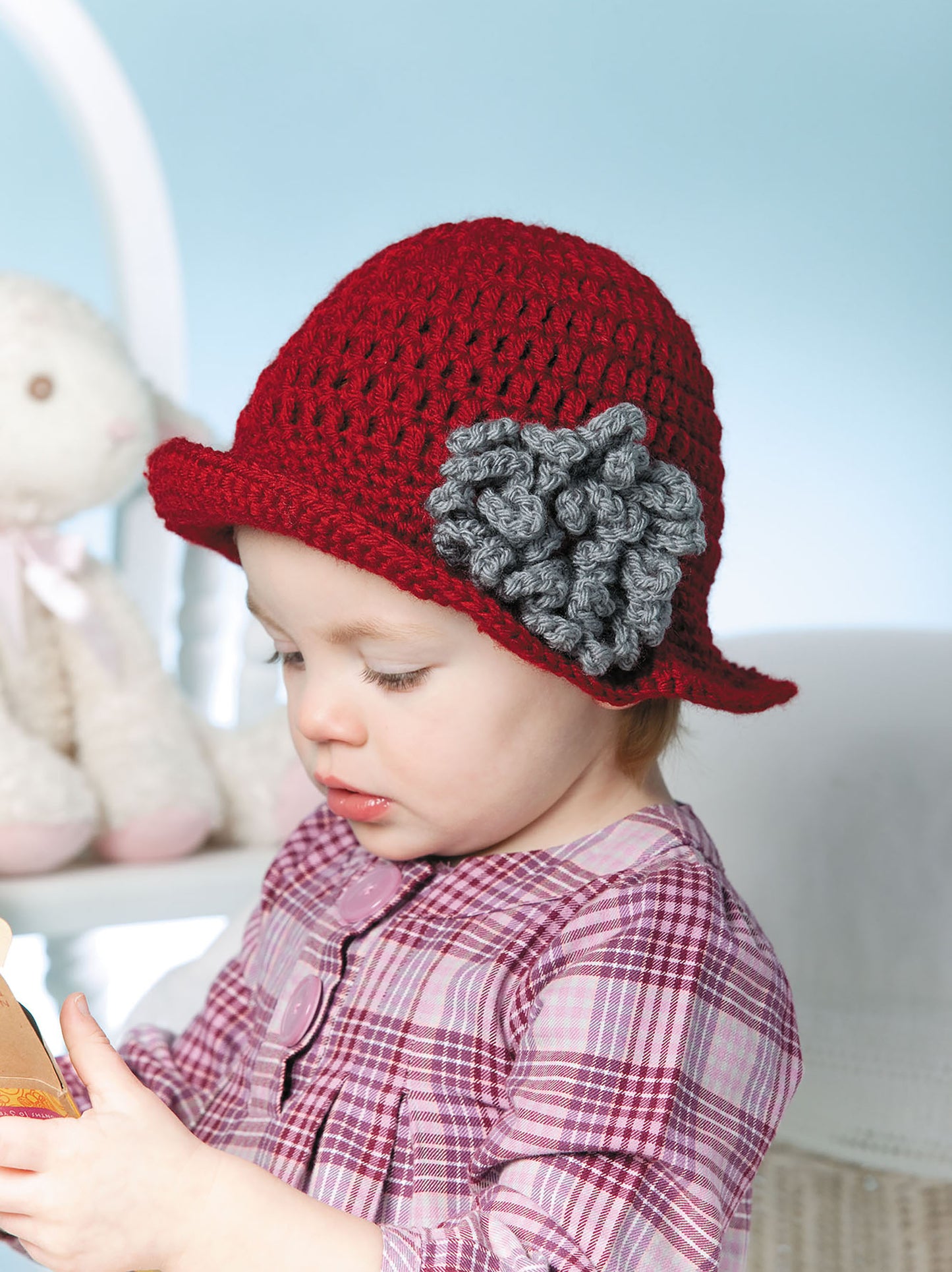 A young child in a red crocheted hat with a gray flower, made using patterns from "Baby Hats—10 Quick and Easy Patterns Written in Three Sizes" by Leisure Arts, sits indoors in a plaid shirt, holding a book with a plush toy blurred in the background.