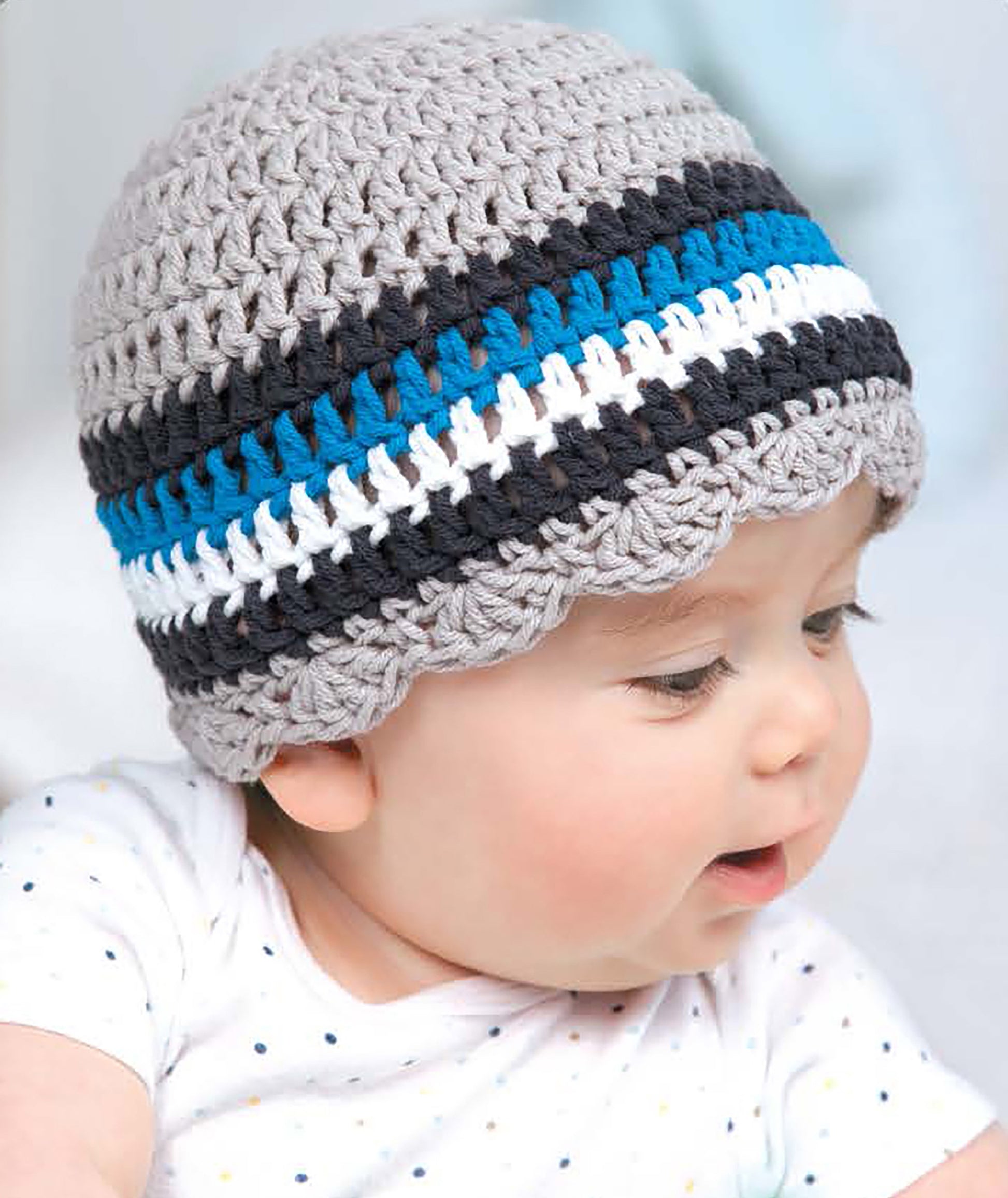 A baby in a striped crocheted hat looks to the side. The hat is from Leisure Arts’ "Baby Hats—10 Quick and Easy Patterns Written in Three Sizes," ideal for beginners or anyone seeking cute newborn crochet patterns. The baby wears a dotted white shirt.