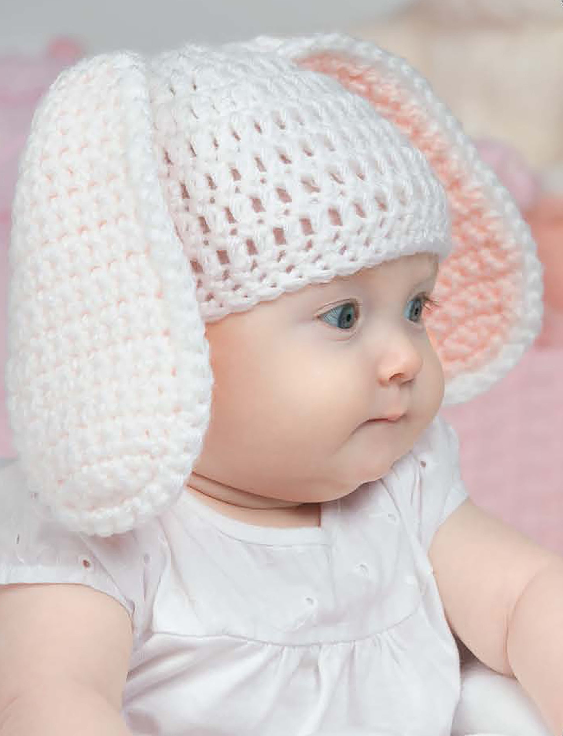 A blue-eyed baby in a white crocheted bunny hat, perfect for beginners, sits sideways in soft pastels. Pattern from "Baby Hats—10 Quick and Easy Patterns Written in Three Sizes" by Leisure Arts.