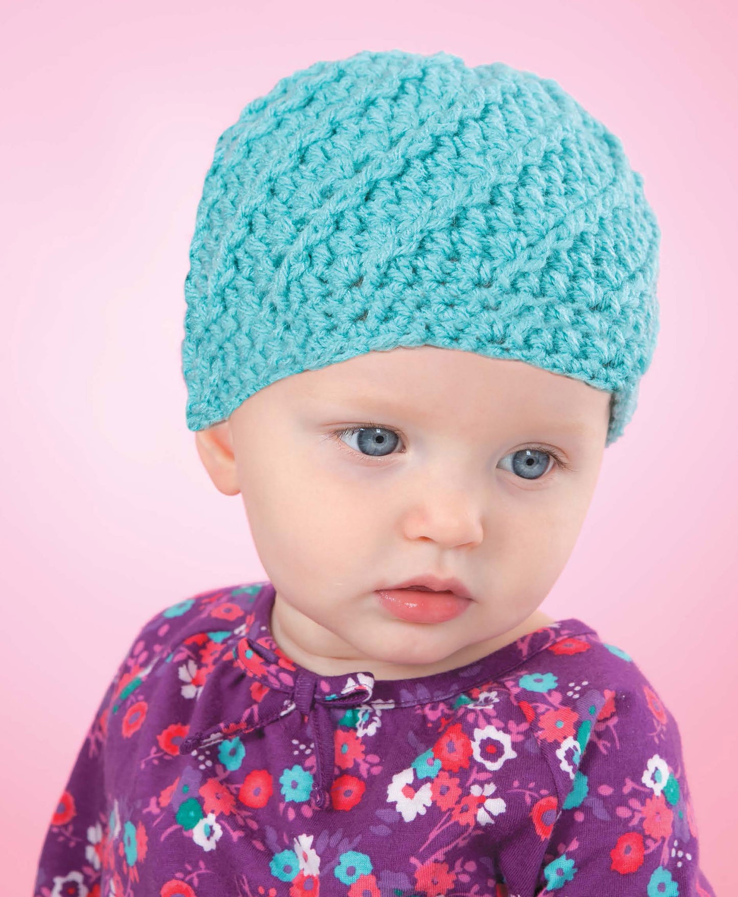 A baby with blue eyes wears a light blue hat inspired by Leisure Arts' "Baby Hats—10 Quick and Easy Patterns Written in Three Sizes," paired with a purple floral top, looking to the side against a soft pink background.