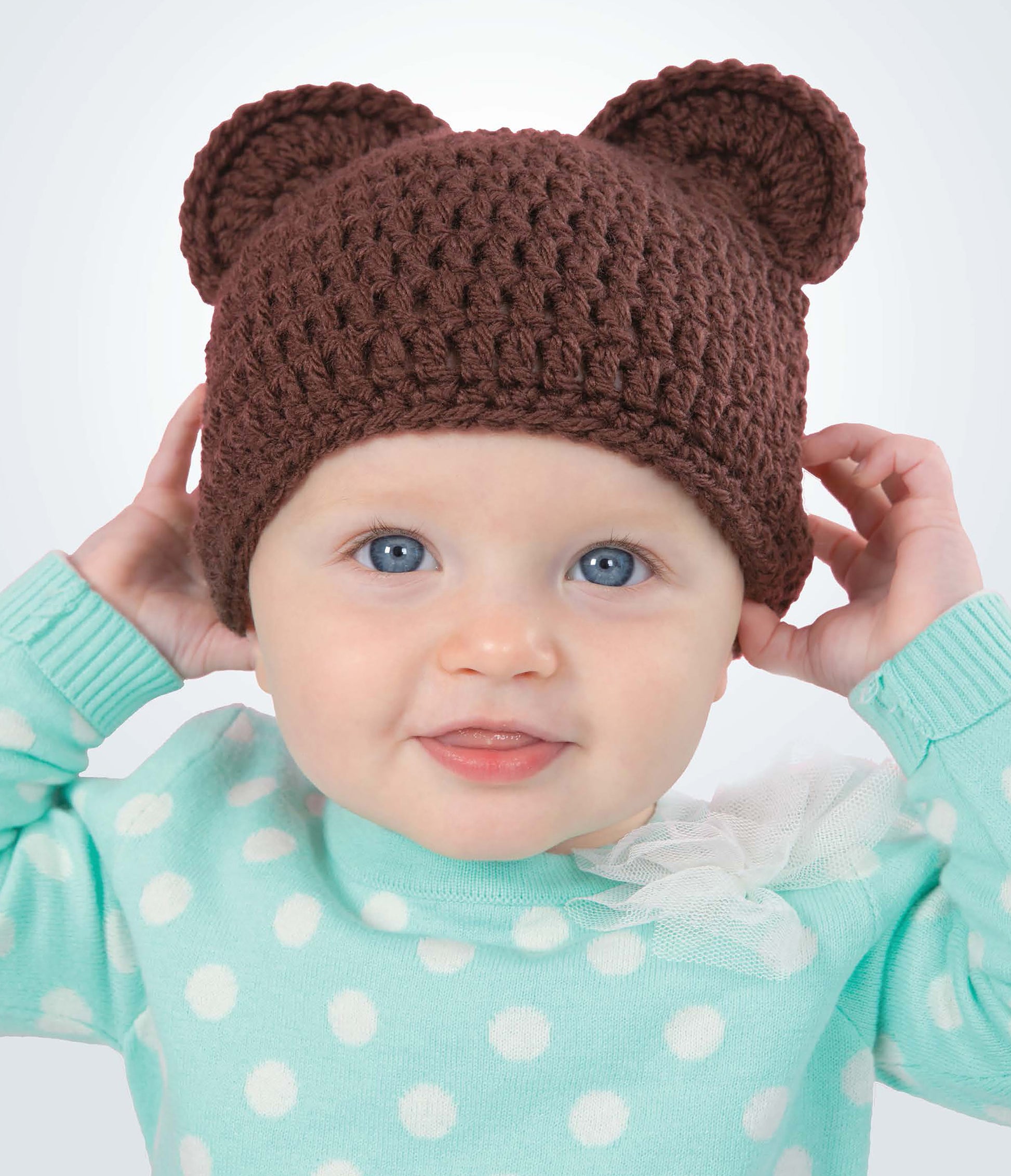 A baby smiles in a brown crocheted bear-ear hat, inspired by "Baby Hats—10 Quick and Easy Patterns Written in Three Sizes" from Leisure Arts.