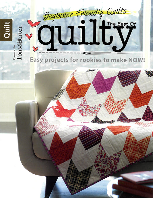 A vibrant quilt with chevron patterns in red, orange, purple, and pink covers a white couch by a sunny window. The magazine, "The Best of Quilty" by Leisure Arts, features beginner-friendly quilts and easy embroidery projects.