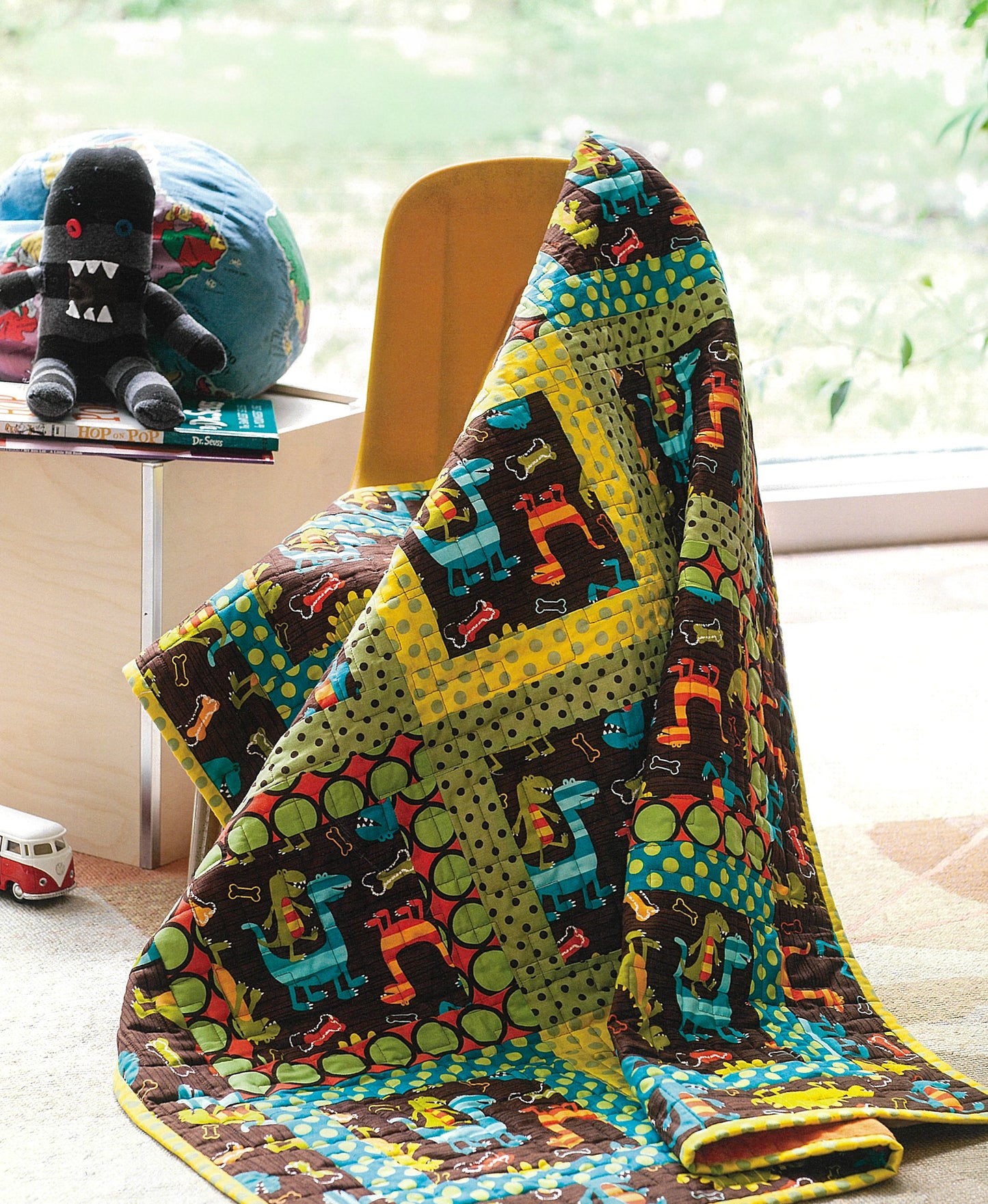 The Best of Quilty by Leisure Arts, a vibrant dinosaur-patterned quilt with illustrated stitches, is draped over a yellow chair near a window, next to a plush toy, toy bus, and globe on a small table.