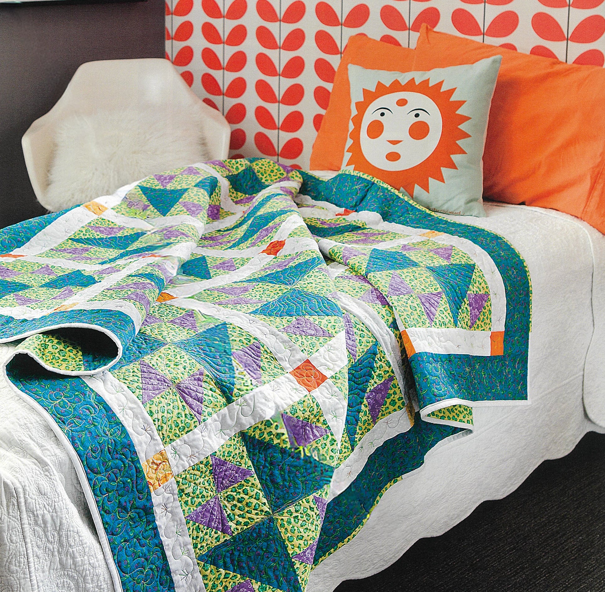 A blue, green, and purple patchwork quilt made with The Best of Quilty by Leisure Arts covers a white bed. An orange sun face pillow sits against a retro wall, while a white chair rests nearby.