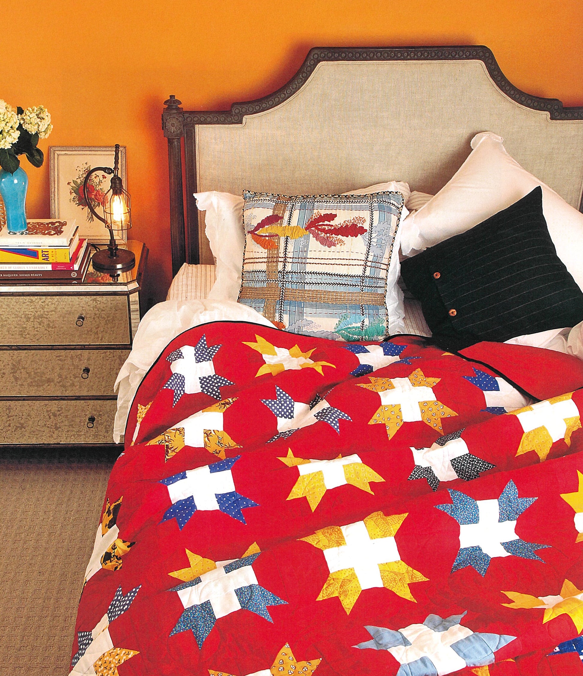 A cozy bed with a beige headboard, colorful pillows, and a vibrant red star-patterned quilt inspired by The Best of Quilty from Leisure Arts. Nearby, a table holds books, a lamp, flowers, and framed art set against an orange wall.