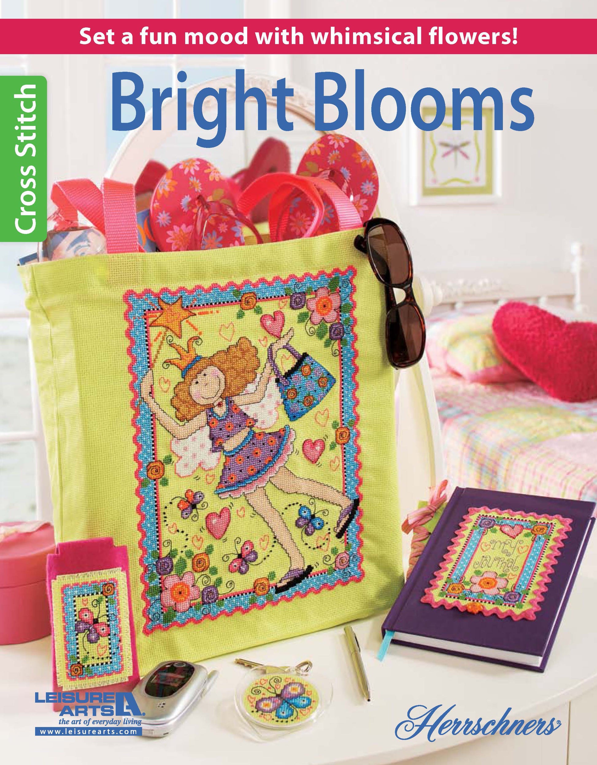 The Bright Blooms Cross Stitch kit by Leisure Arts features playful flower designs on a green tote, matching notebook, glasses case, and keychain. Sewing supplies and pink decor complete the look. Text: Bright Blooms Cross Stitch.