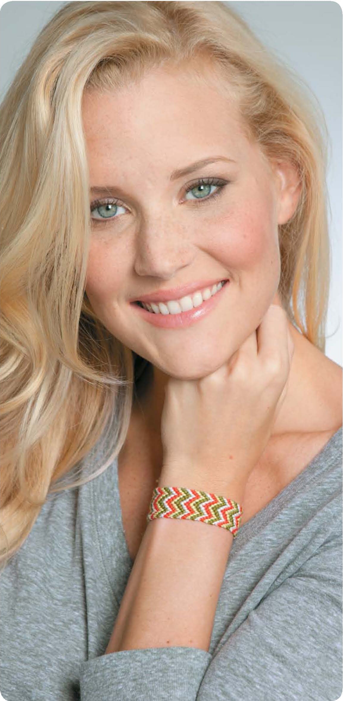 A woman with long blonde hair and green eyes smiles at the camera, wearing a light grey top and displaying colorful bracelets made with the "Friendship Bracelets Beyond the Basics Digital Download" by Leisure Arts.