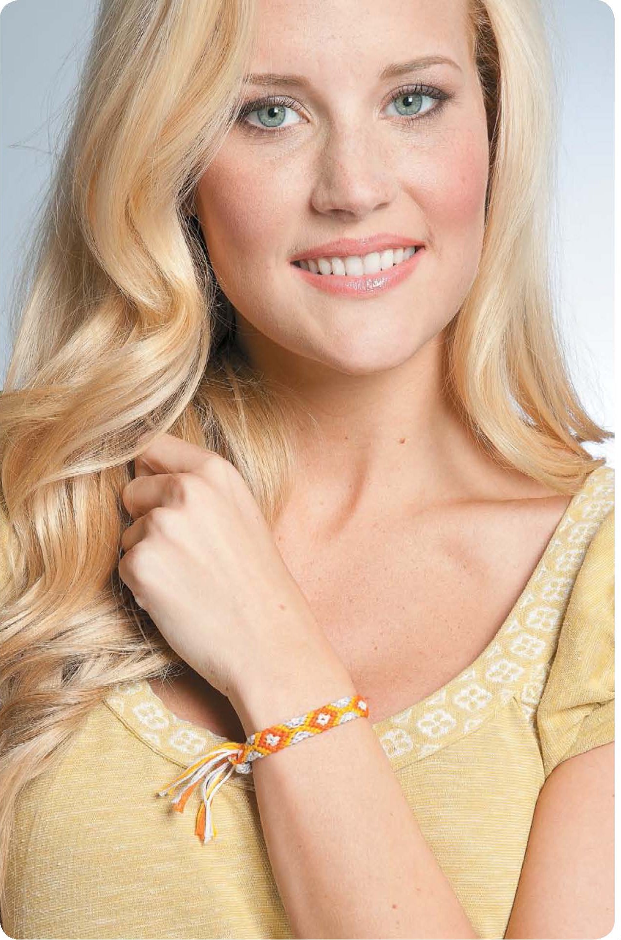 A smiling woman with long blonde hair models a yellow and white friendship bracelet from Leisure Arts’ "Friendship Bracelets Beyond the Basics" digital download, wearing a yellow top and looking at the camera.