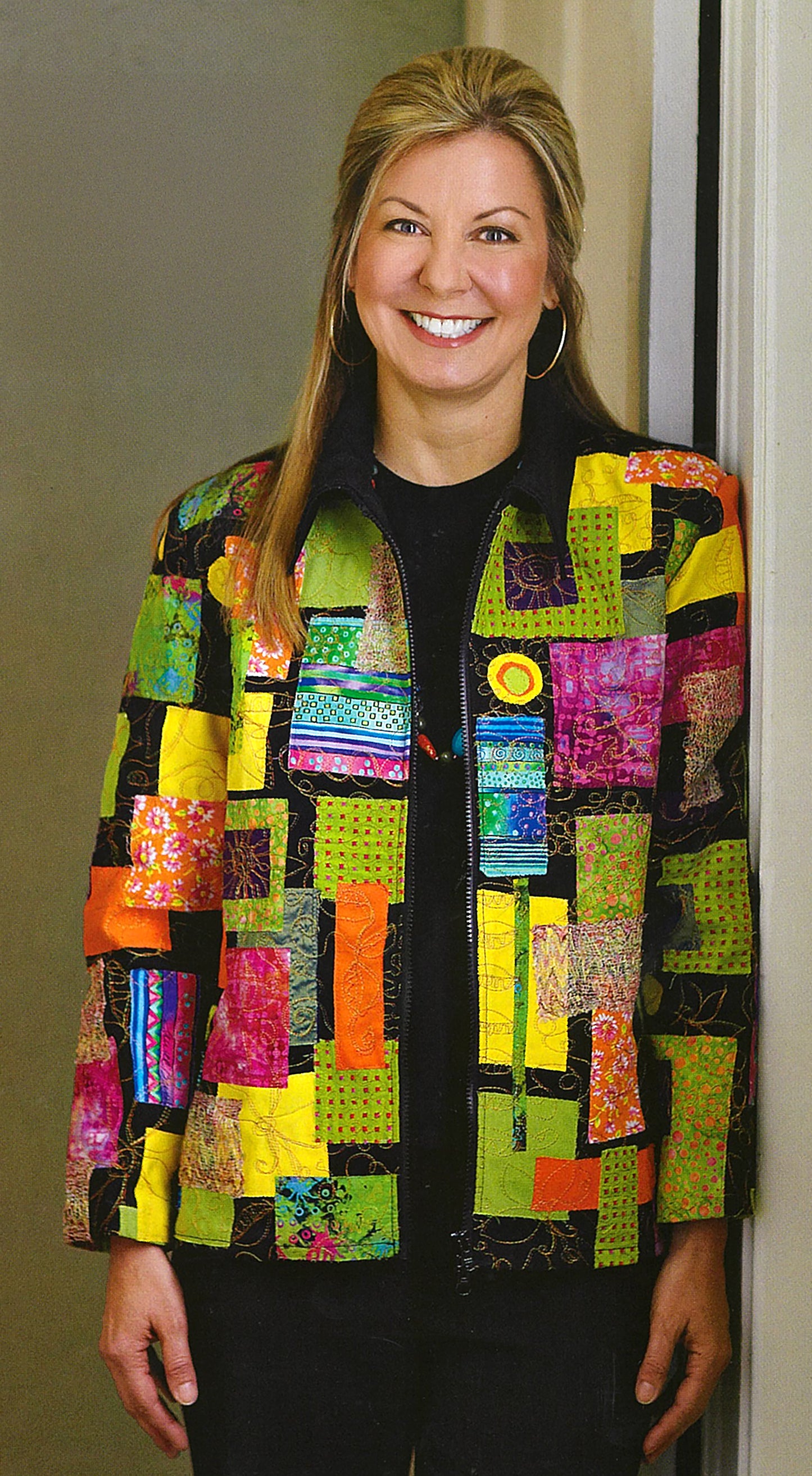 A smiling woman with long blonde hair stands by a doorway, wearing a vibrant patchwork jacket made using chic techniques from Sew Basics & Beyond: Simple Embellishments by Leisure Arts, featuring colorful green, yellow, orange, pink, and black fabric pieces.
