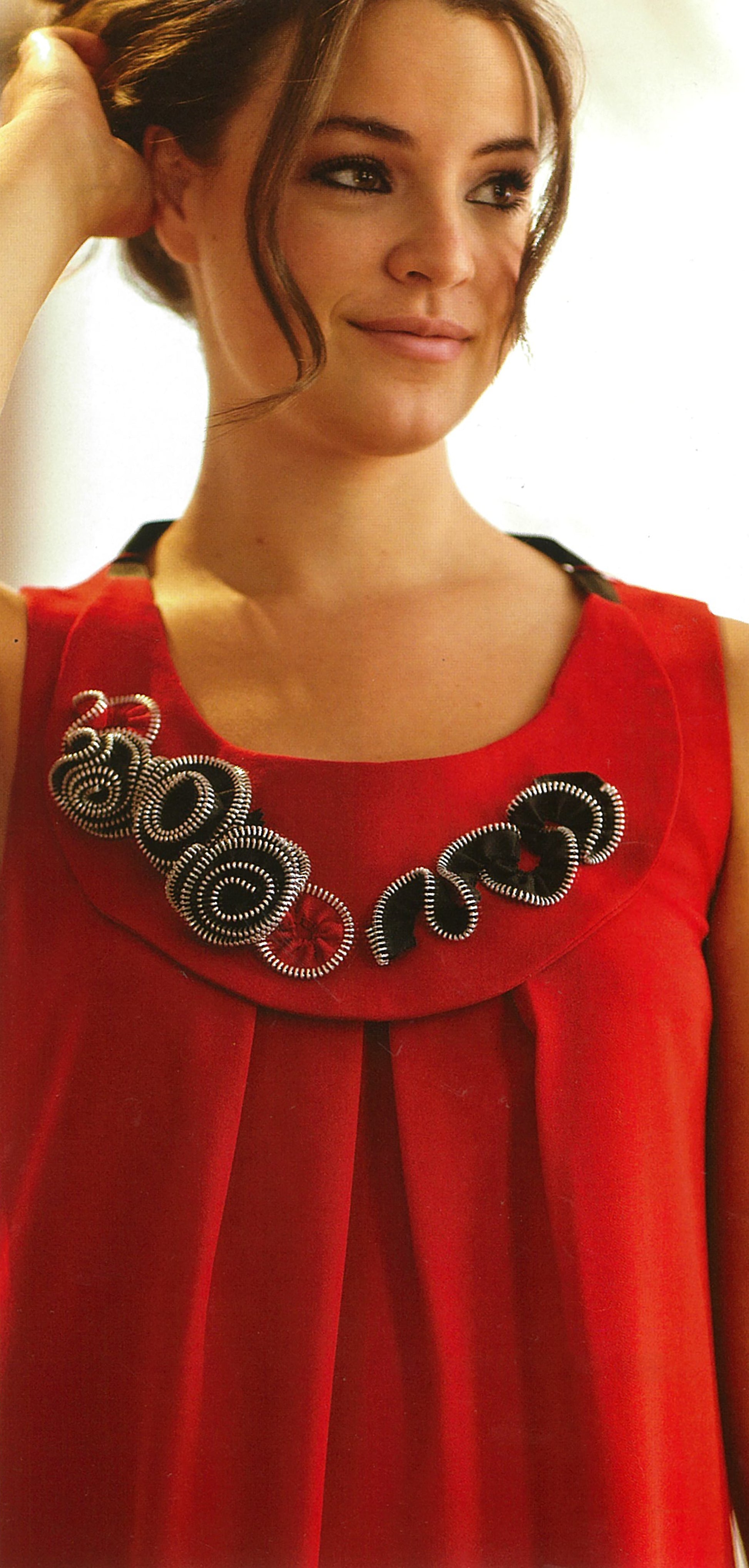 A woman in a sleeveless red dress with a spiral and wavy black, gray, and red necklace—her light brown hair styled up—smiles softly to the side, showcasing chic techniques from Sew Basics & Beyond: Simple Embellishments by Leisure Arts.