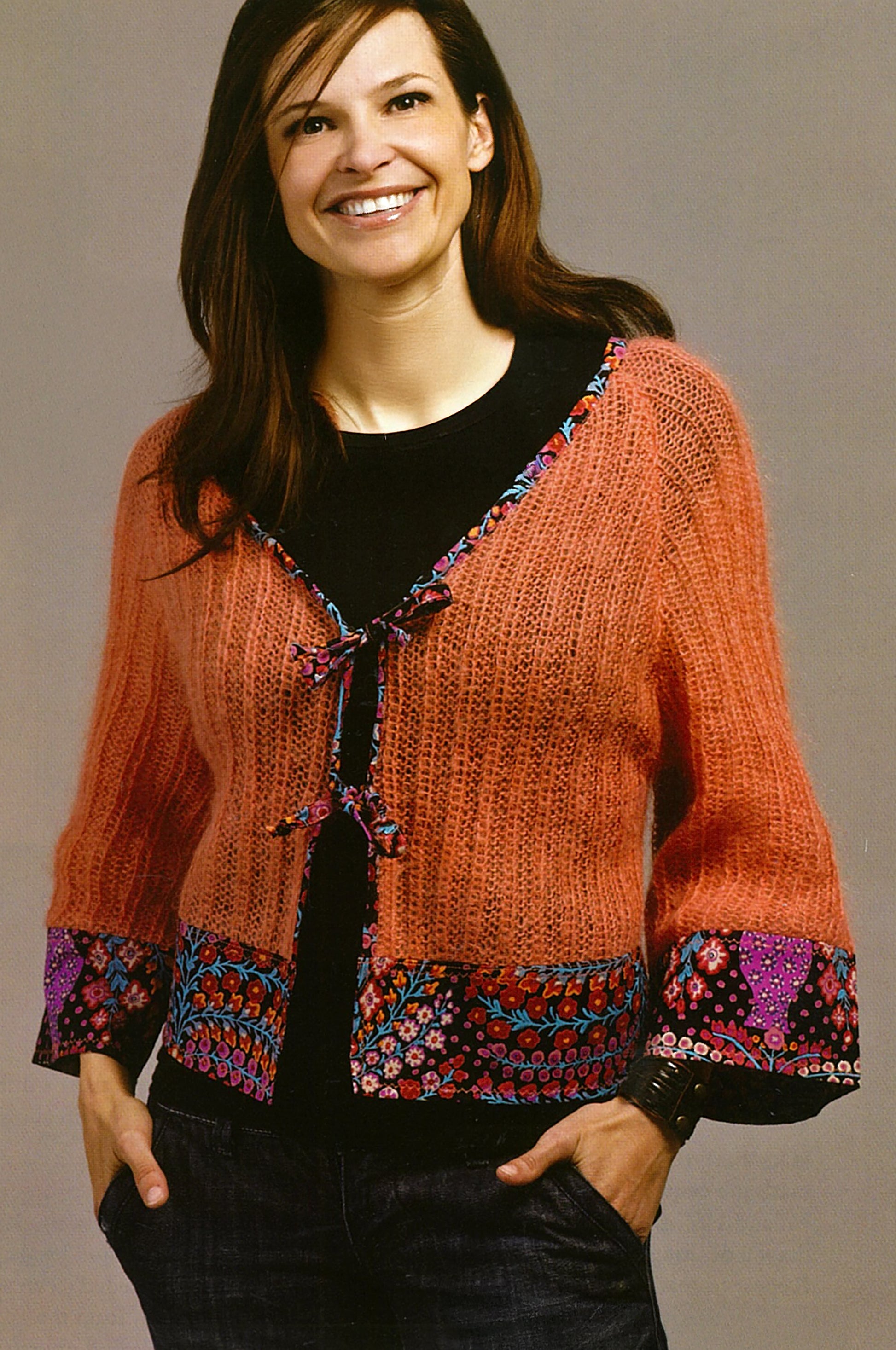 A woman with long brown hair smiles in a rust knit cardigan with floral trim, styled over a black top and jeans. She showcases inspiration from Sew Basics & Beyond: Simple Embellishments by Leisure Arts against a plain background.