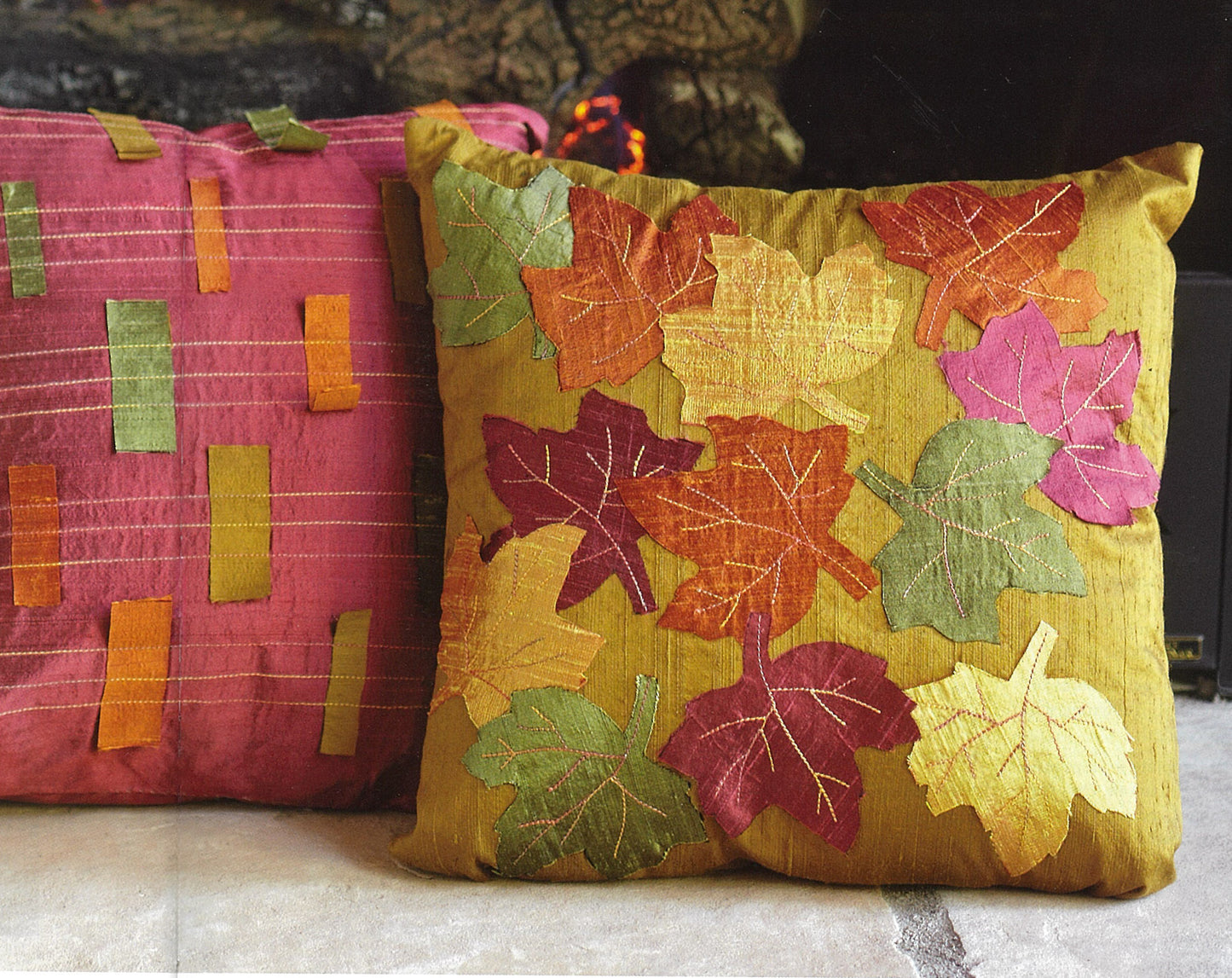Add plush comfort and style to your home with Leisure Arts' "Pillows—Accent Your Home." One features gold fabric with embroidered autumn leaves, the other pink-red with green and orange patches—both showcase creative, textured handmade designs.