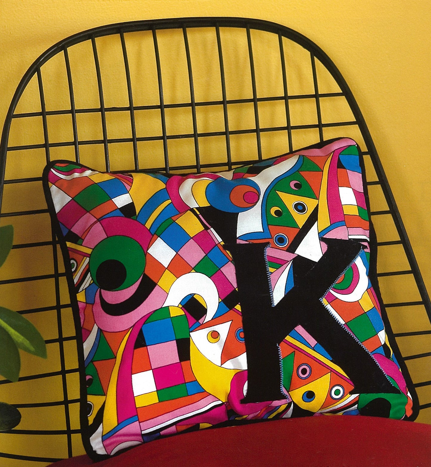The Leisure Arts Pillows—Accent Your Home with Plush Comfort & Style! square pillow features a vibrant abstract pattern and bold black "K," displayed on a black wire chair by a yellow wall with a plant leaf peeking in the corner.