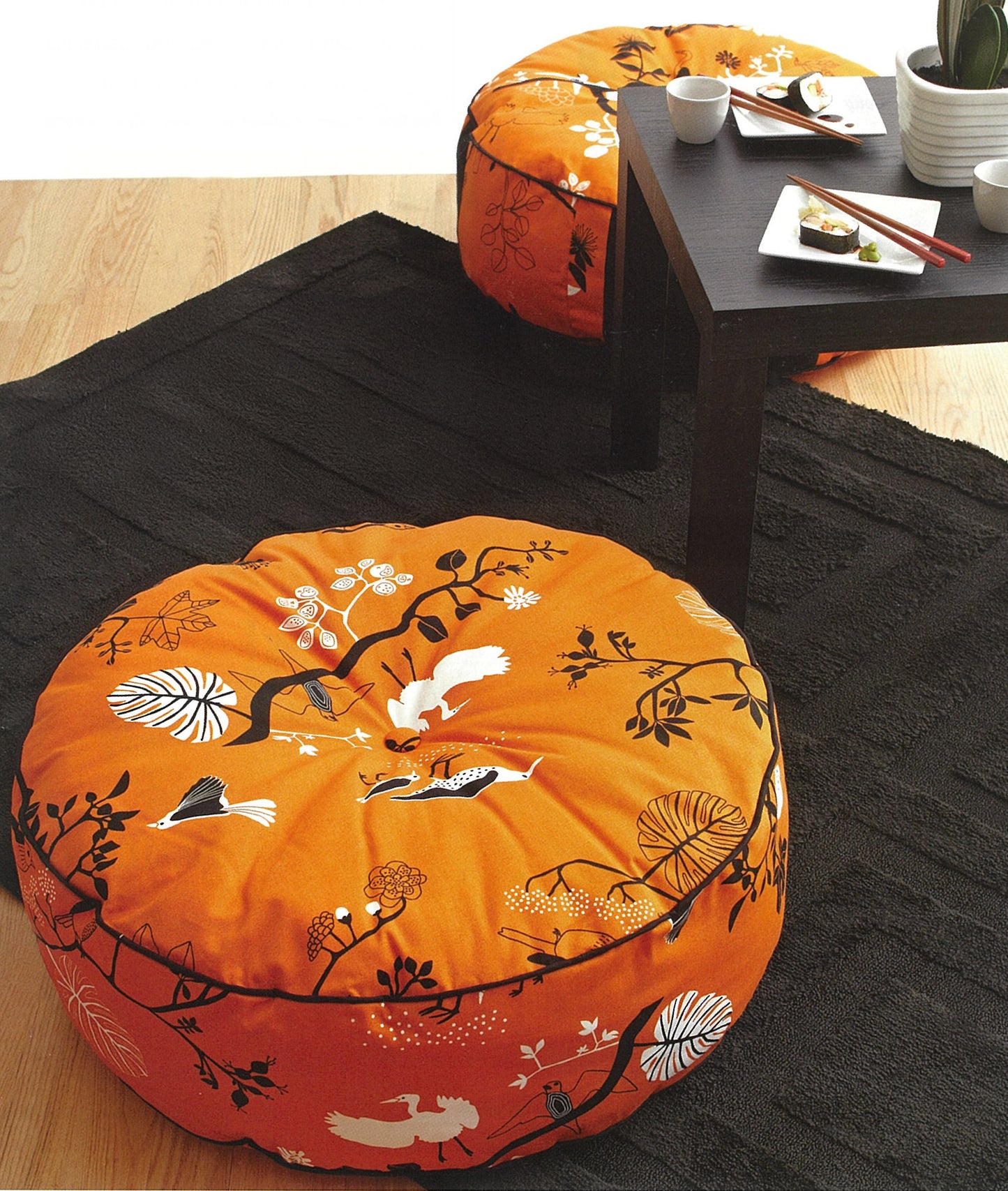 Two round, orange Pillows—Accent Your Home with Plush Comfort & Style! from Leisure Arts, featuring floral and animal prints, rest on a black rug by a low table set with white dishes and sushi on a wooden floor.