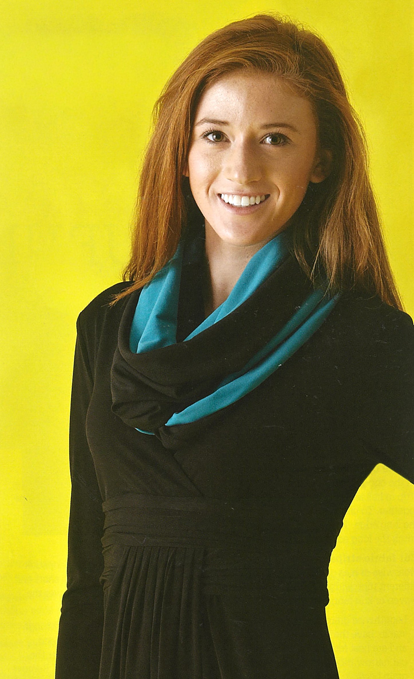 A woman with long auburn hair, in a black dress and a blue-black scarf she made using Leisure Arts’ Sew it All: Quick Projects, smiles against a bright yellow background.