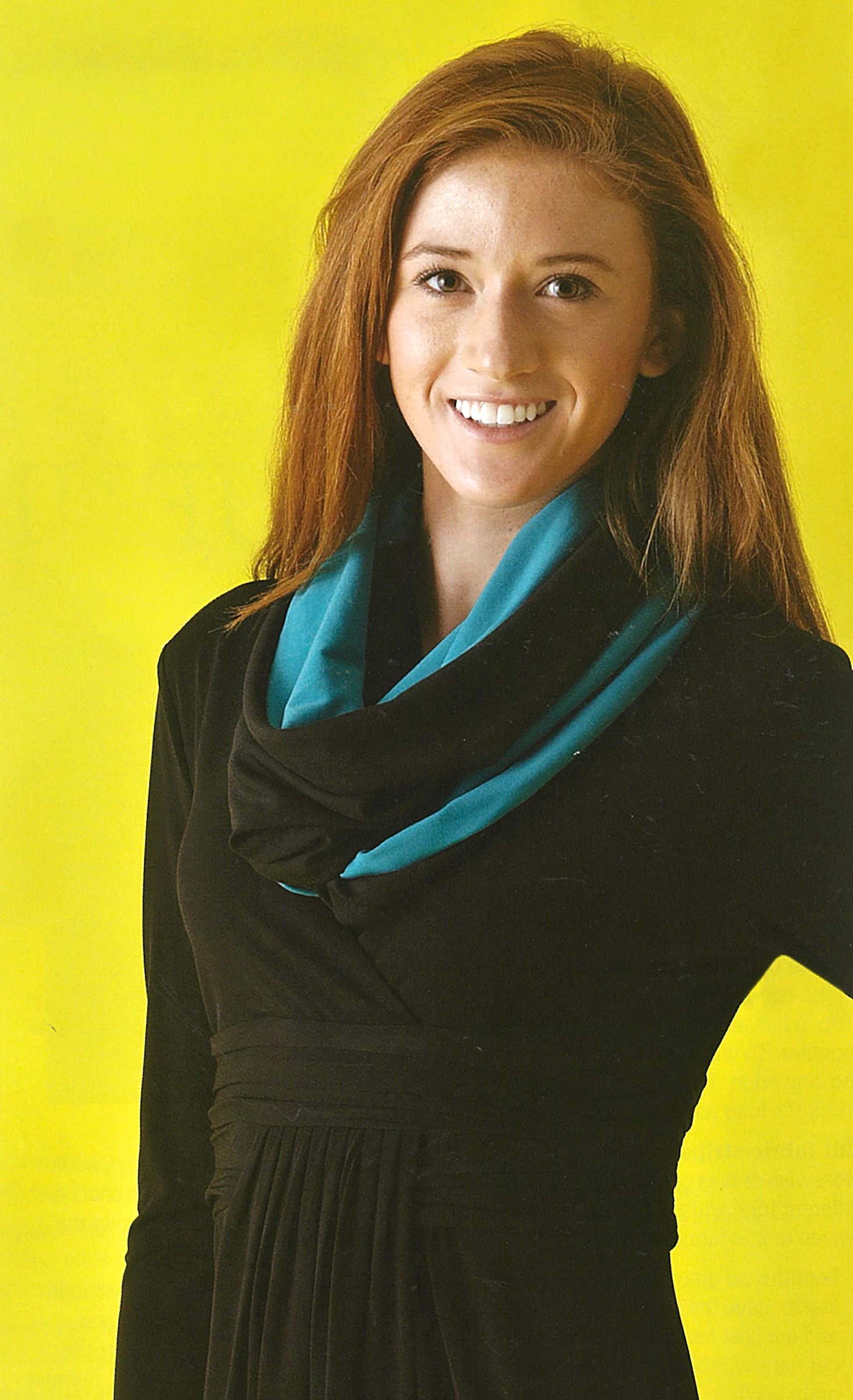 A woman with long auburn hair, in a black dress and a blue-black scarf she made using Leisure Arts’ Sew it All: Quick Projects, smiles against a bright yellow background.