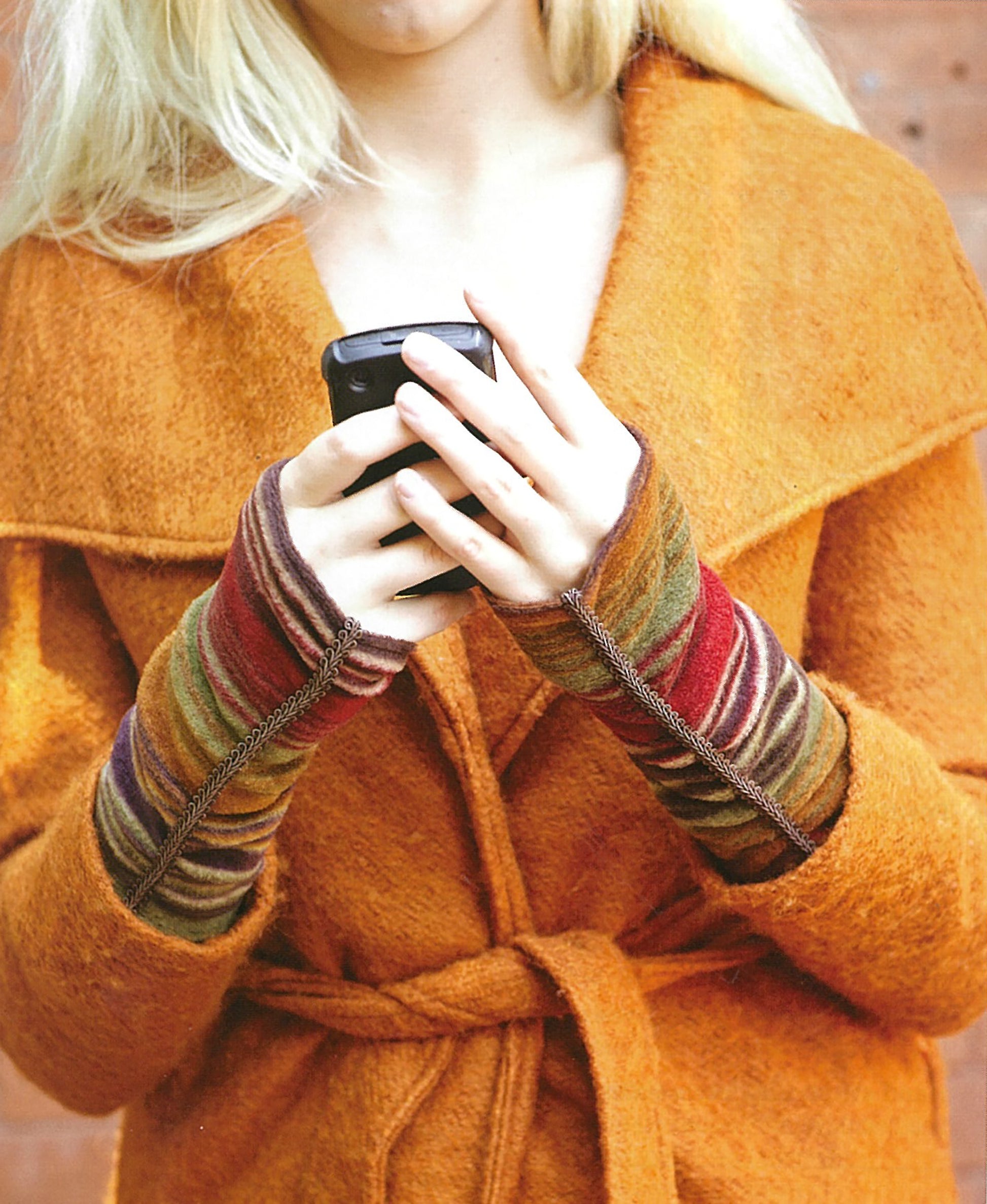 A person in a bright orange coat and colorful, striped fingerless gloves—ideal for newbies using Leisure Arts' Sew it All: Quick Projects—is holding a smartphone, with their face partially out of frame.