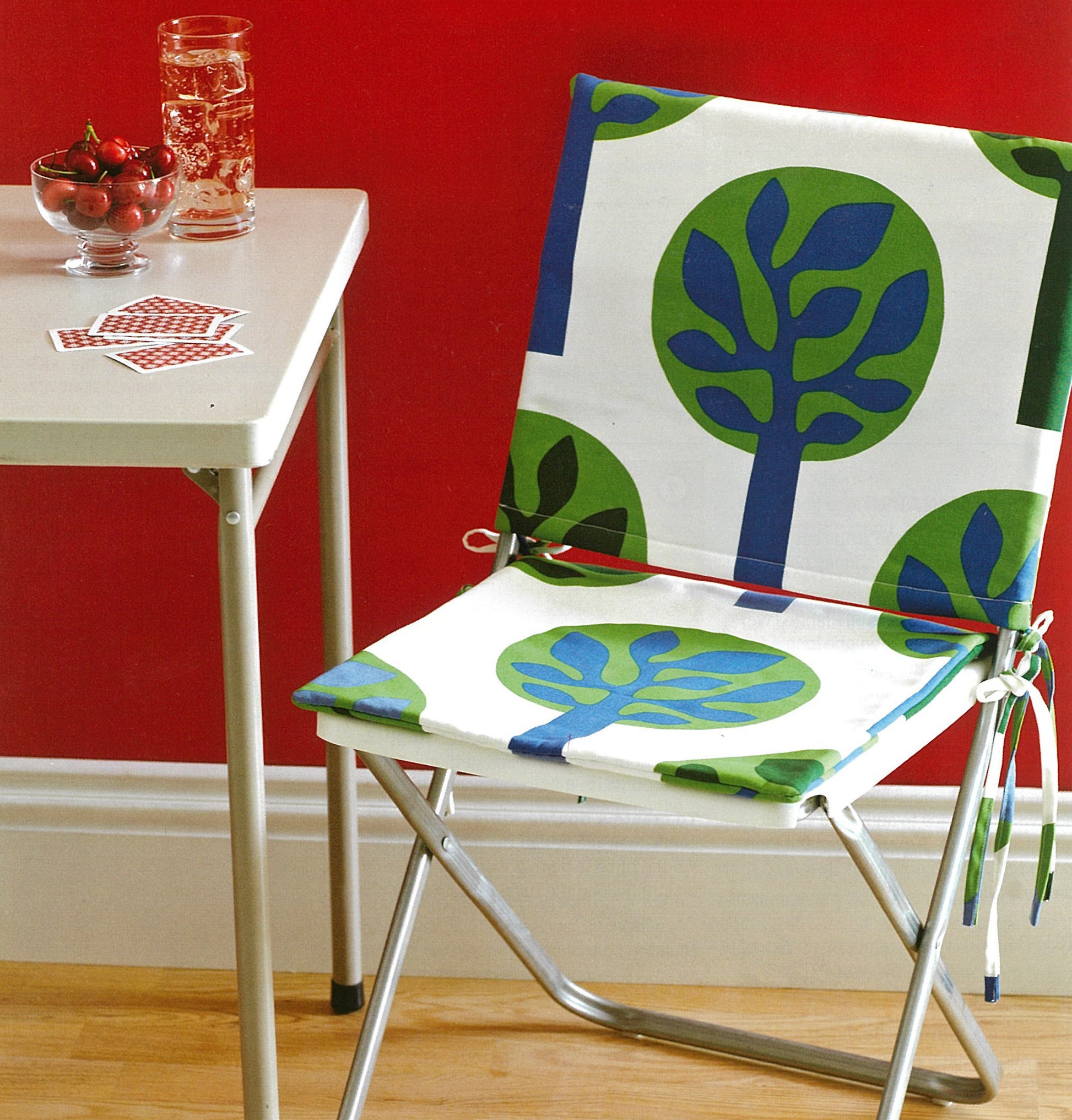 A folding chair with a green and blue tree print, perfect for planning beginner projects from Leisure Arts' Sew it All: Quick Projects, sits by a small table with water, cherries, and cards, set against a red wall and wood floor.