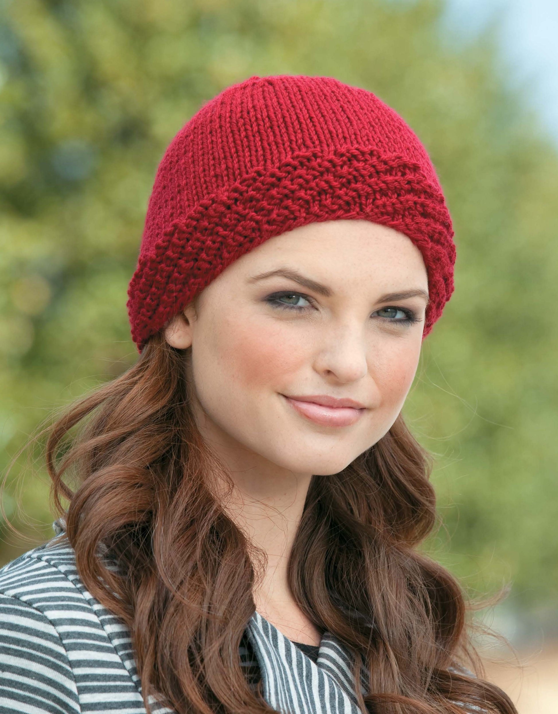 A woman with long, wavy brown hair wears a red knit hat made using Leisure Arts’ Beginner's Guide to Knitting in the Round, paired with a black-and-white striped top while standing outdoors against blurred trees.
