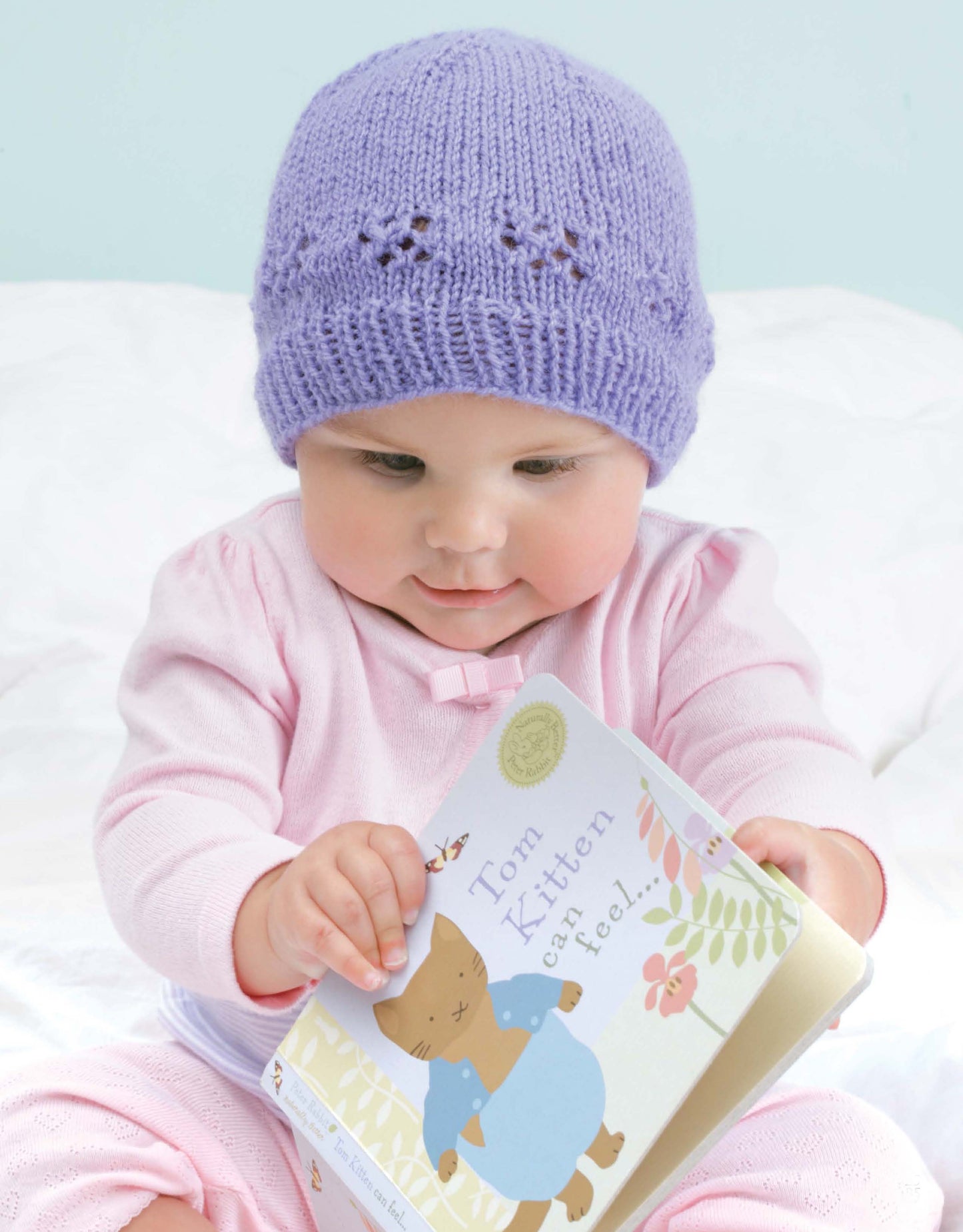 A baby in a lavender knit hat—a great beginner project from the Leisure Arts Beginner's Guide to Knitting in the Round—looks down while holding a children's book with Tom Kitten on the cover.
