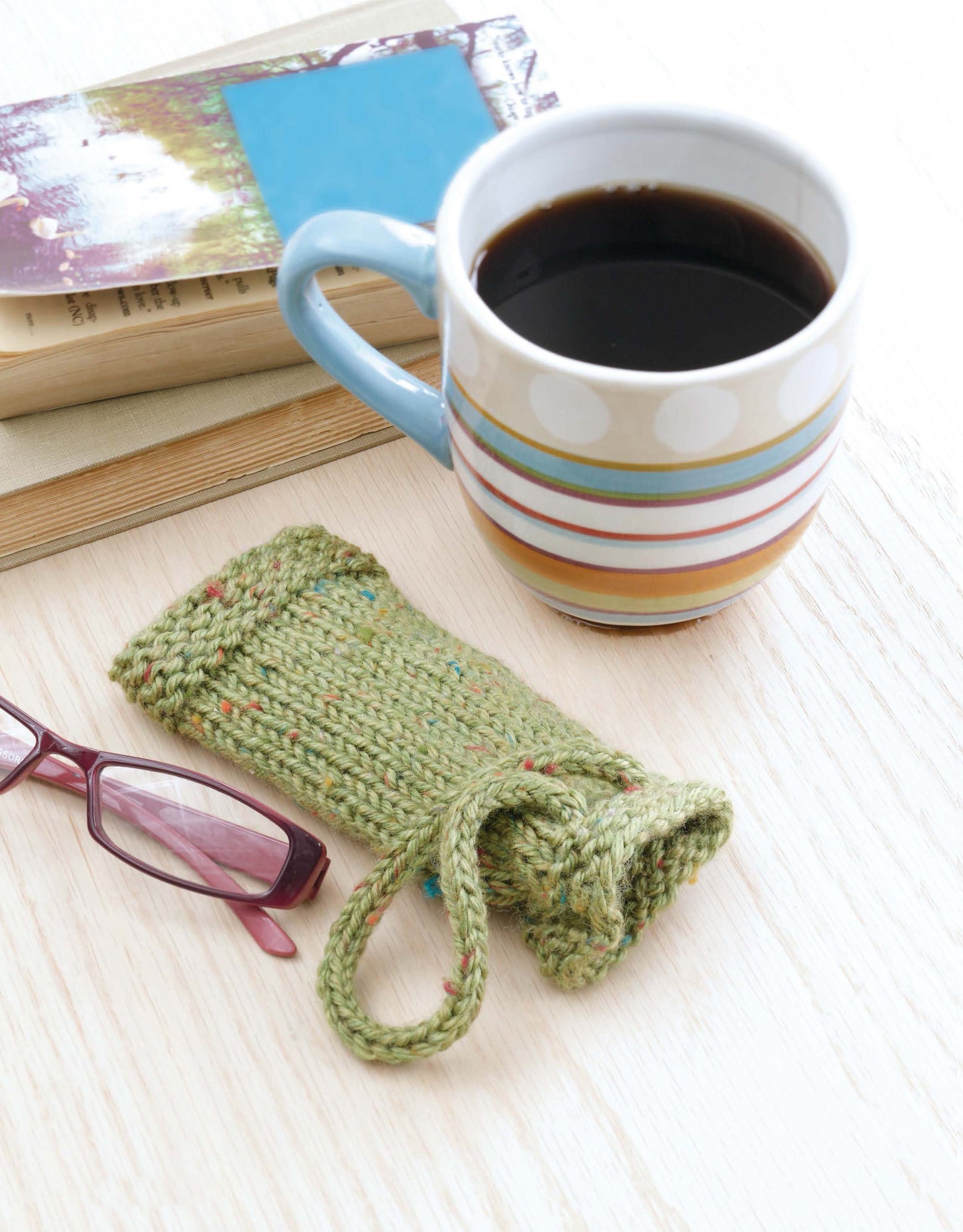 A mug of black coffee sits on a desk beside the Beginner's Guide to Knitting in the Round by Leisure Arts, with red glasses and a green knitted case—ideal inspiration for learning circular knitting techniques.