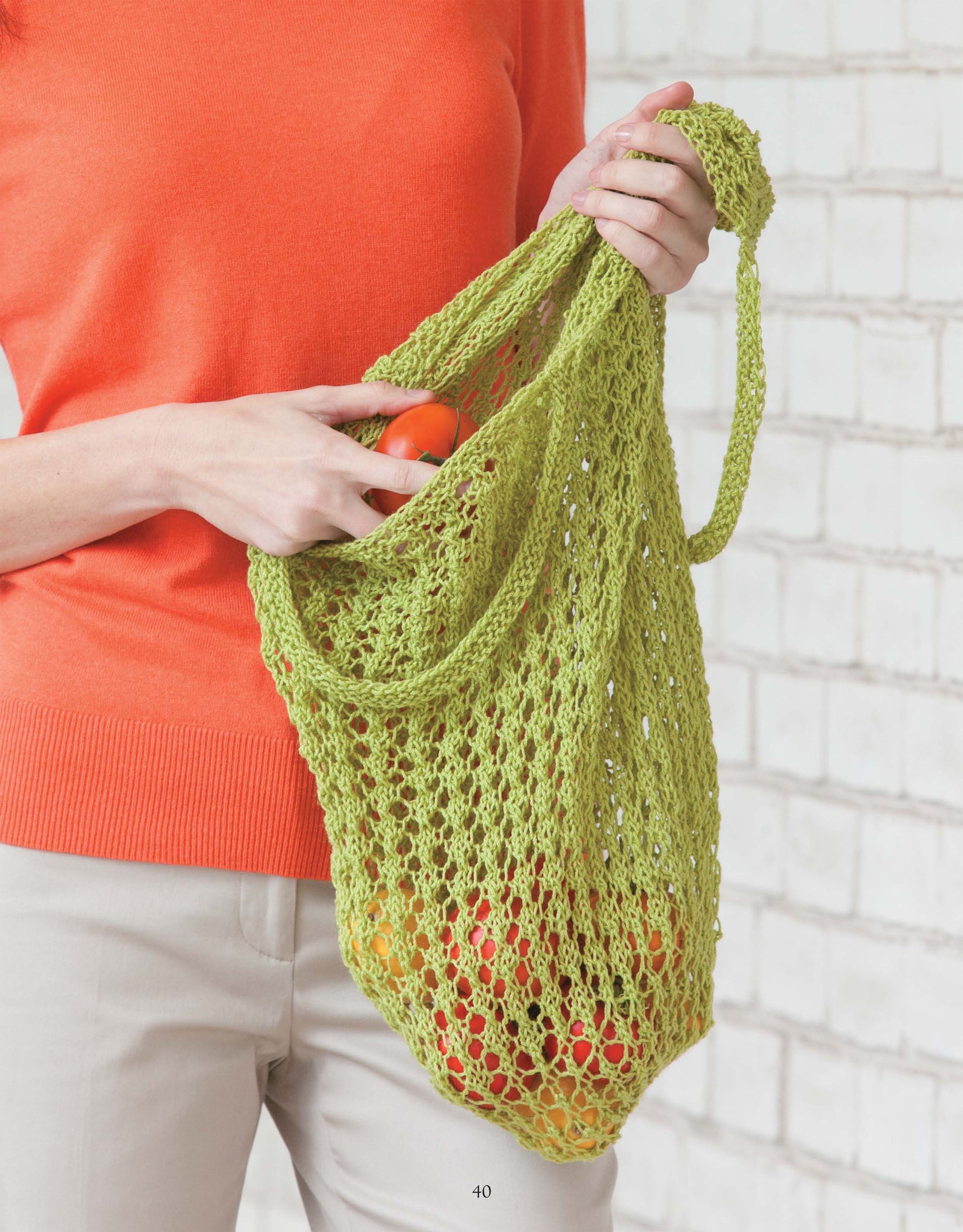 A person in a bright orange sweater and beige pants holds a green, crocheted mesh bag of tomatoes—an inspiring look for anyone exploring lace patterns in the Beginner's Guide to Lace Knitting by Leisure Arts, posed before a white brick wall.