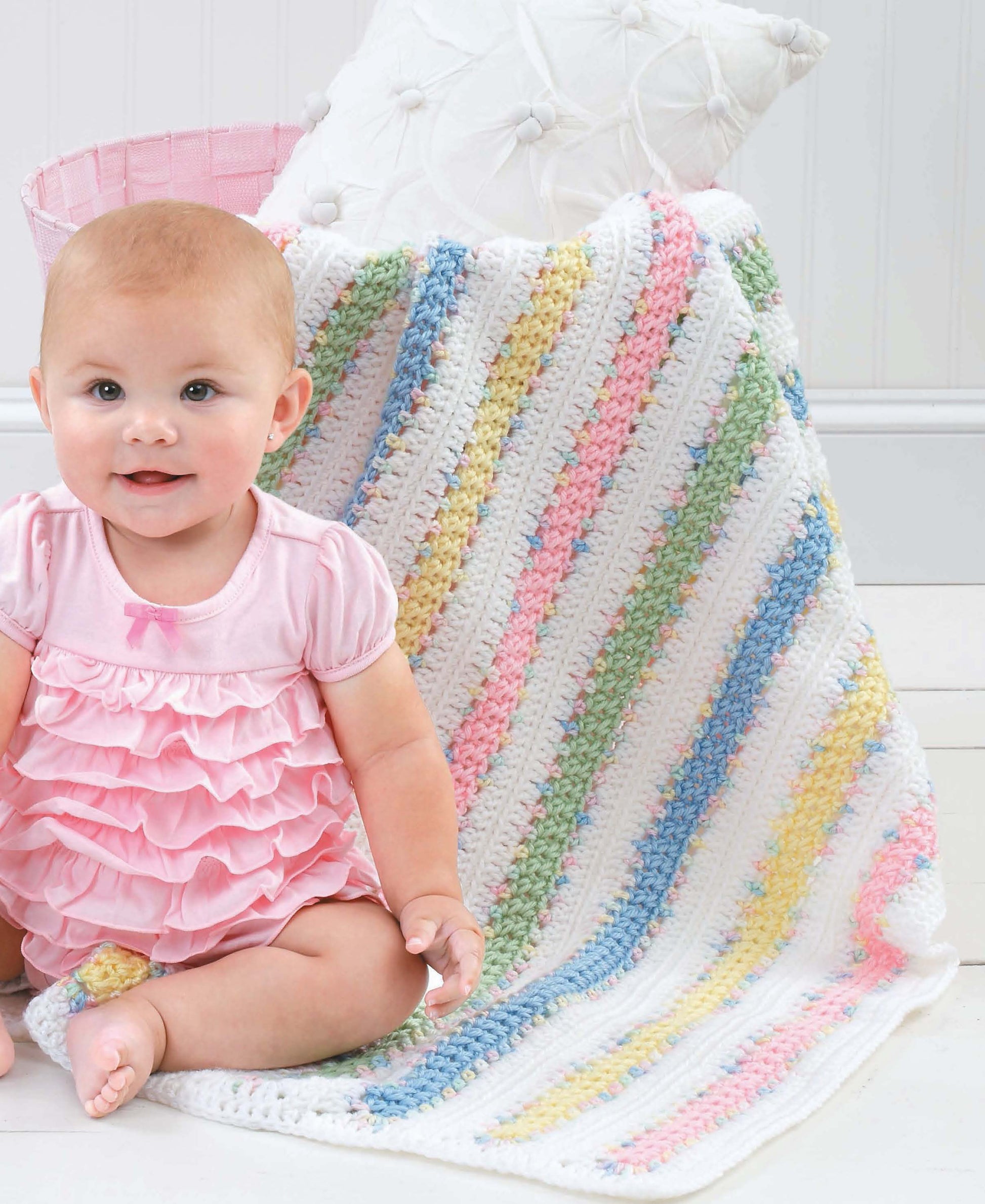 A smiling baby in a pink ruffled dress sits by a pastel-striped Crochet Blankets for Every Baby by Leisure Arts, made with worsted weight yarn and draped over a white cushion.