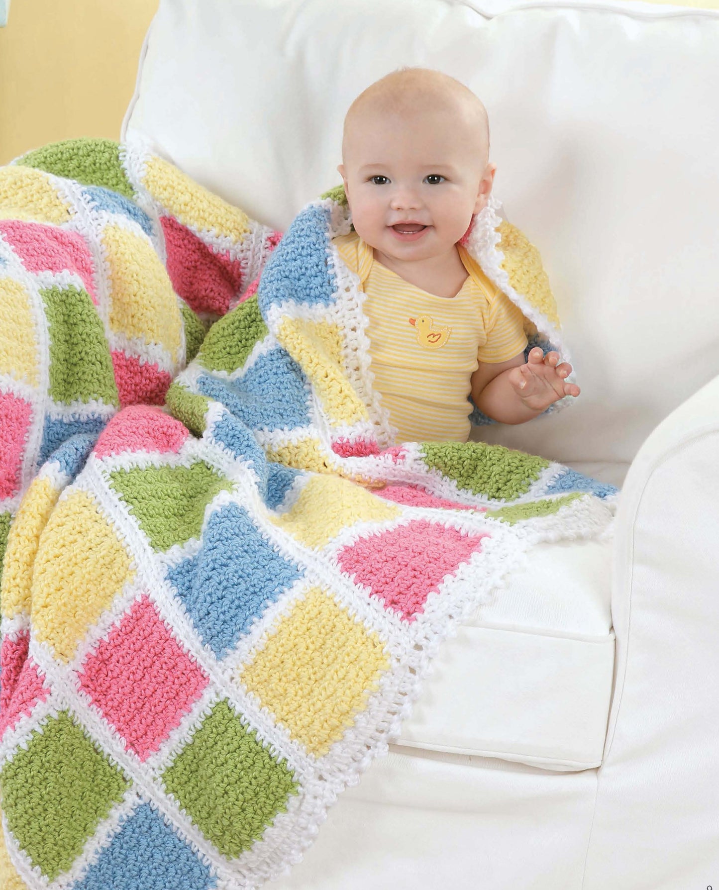 A smiling baby in a yellow onesie sits on a white couch, wrapped in Leisure Arts’ "Crochet Blankets for Every Baby," featuring colorful blue, green, pink, and yellow squares bordered in white.