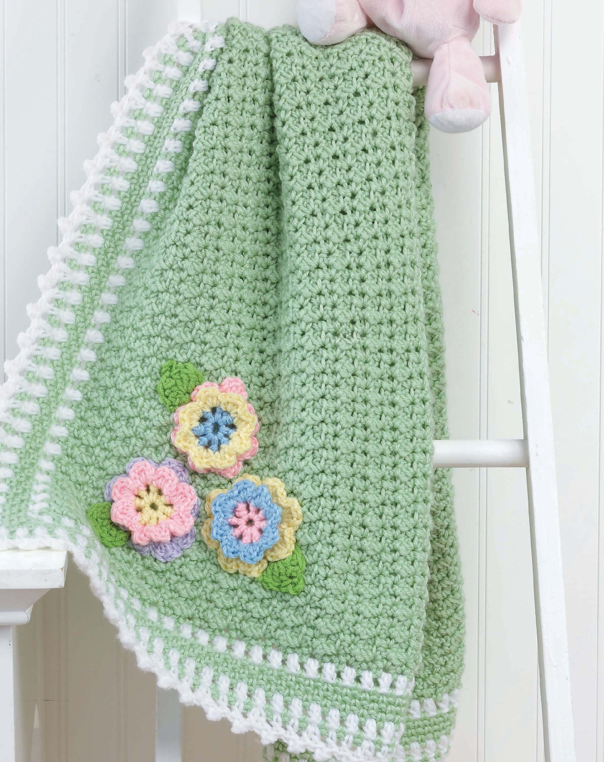 Crochet Blankets for Every Baby" by Leisure Arts features a soft green worsted weight blanket with a white border and three colorful flowers, displayed on a ladder with a pink stuffed animal—perfect inspiration for your next baby project.