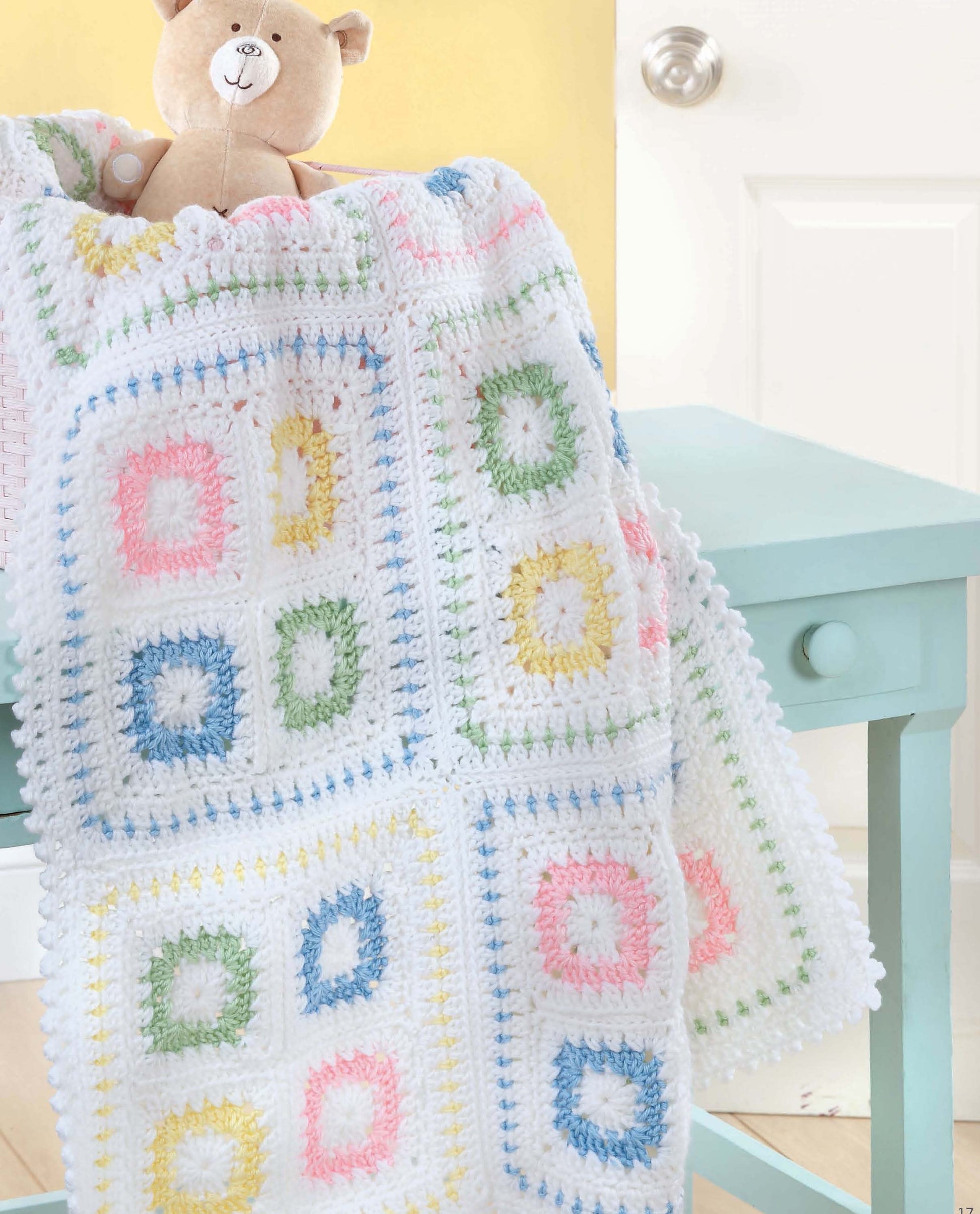 The Crochet Blankets for Every Baby by Leisure Arts, featuring pastel pink, blue, green, and yellow square patterns in worsted weight yarn, drapes over a light blue table with a tan teddy bear peeking out and a white door in the background.