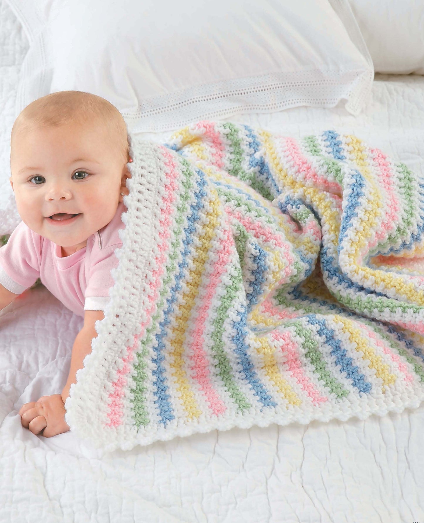 A baby in pink lies on a white bed, partially covered with a pastel striped blanket made from Leisure Arts' Crochet Blankets for Every Baby, crafted in worsted weight yarn of white, pink, yellow, green, and blue.