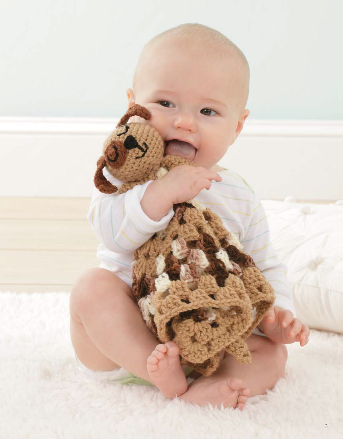 A baby sits on a white rug, smiling and chewing on an Animal Lovie Blanket by Leisure Arts, while wearing a light striped onesie.