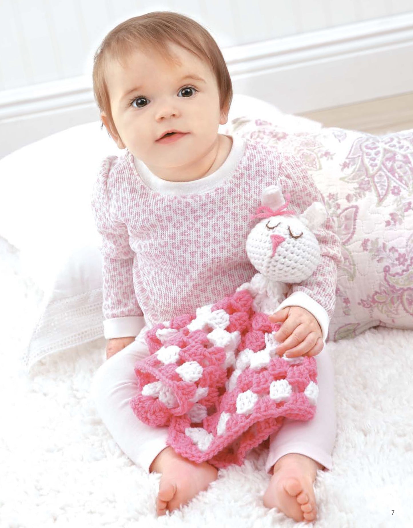 A baby, dressed in pink and white, sits on a fluffy rug holding a Leisure Arts Animal Lovie Blanket, with a patterned pillow behind them. The crochet blanket is inspired by an easy beginner pattern.