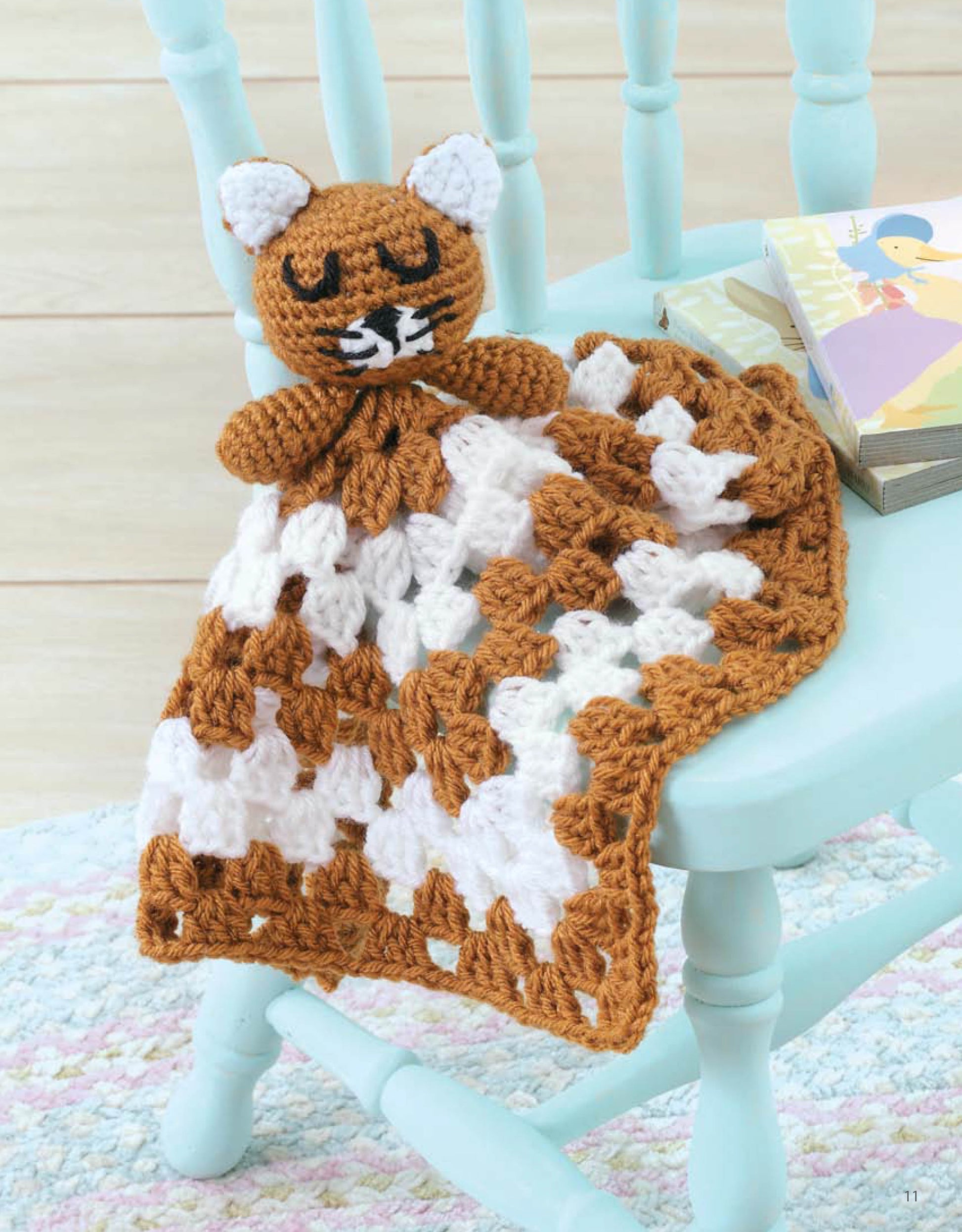 The Leisure Arts Animal Lovie Blankets, featuring a crocheted cat with a brown and white granny square blanket, is draped over a light blue chair beside a book and small stack of items.