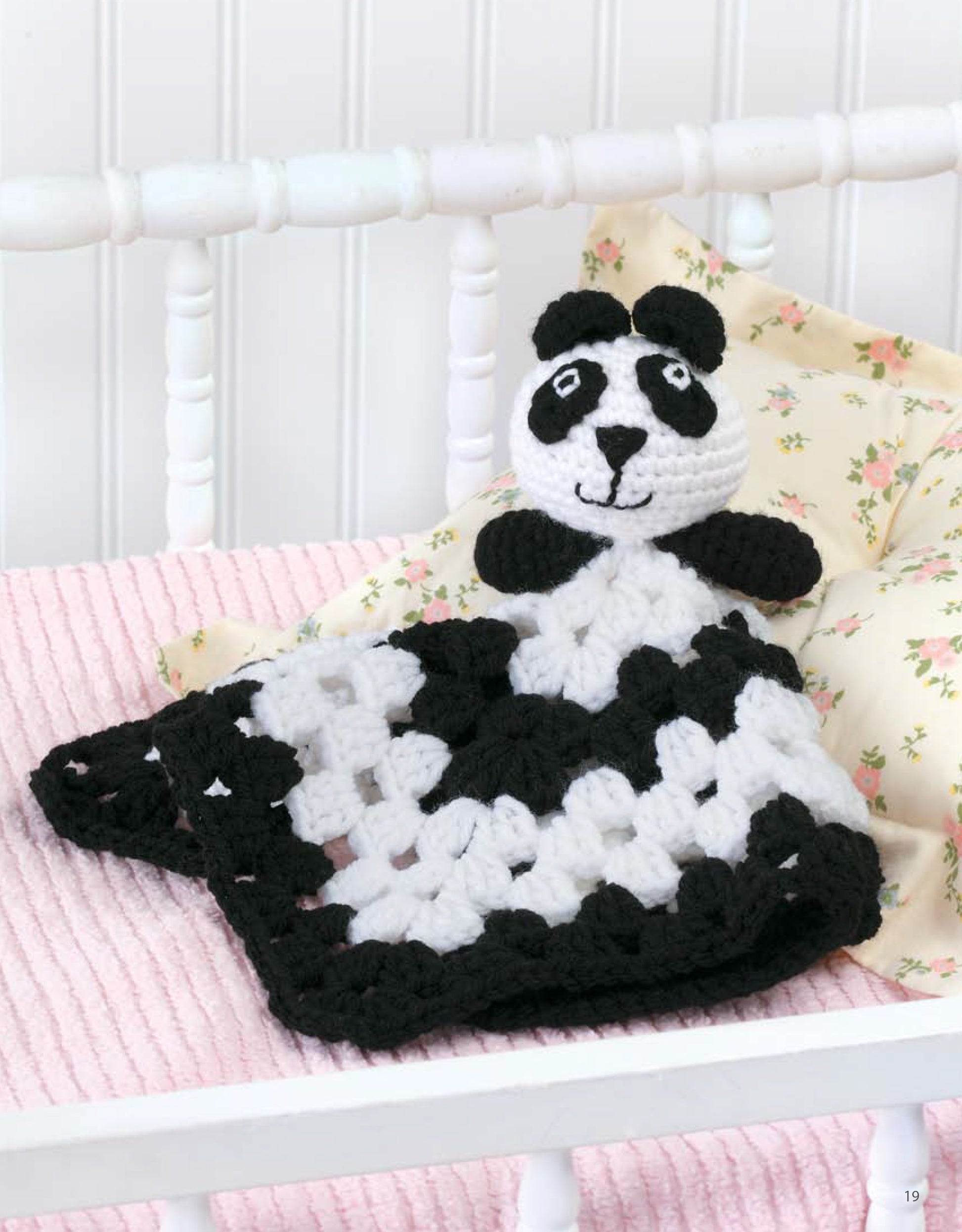 The Leisure Arts Animal Lovie Blankets features a crocheted panda head and arms atop a pink striped blanket, displayed in a white crib before a floral pillow—perfect as a crochet baby blanket or beginner-friendly project.