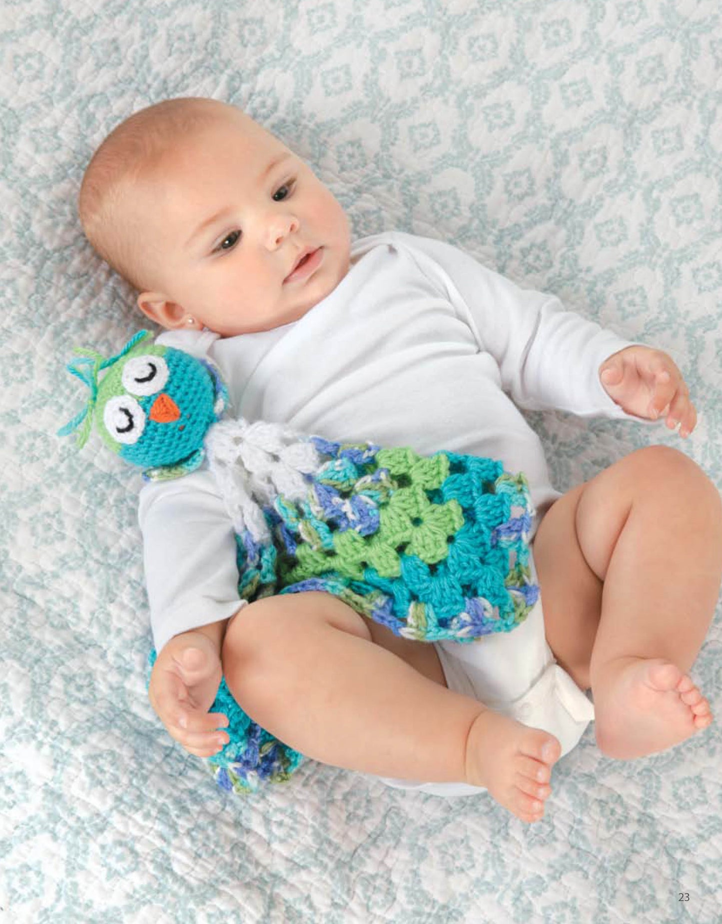 A baby in a white onesie lies on a light blue granny square blanket, holding a Leisure Arts Animal Lovie Blanket—a colorful crocheted owl comforter in blue, green, and white.