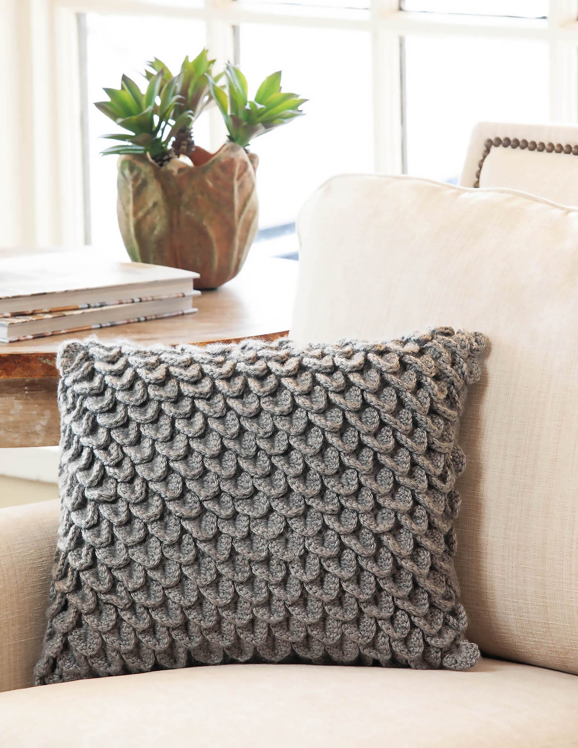 A textured gray pillow featuring a fish-scale pattern, inspired by Leisure Arts' "Beginner's Guide to Crocodile Stitch," rests on a beige armchair. Nearby, a potted plant and stacked books sit on a wooden table by the window.