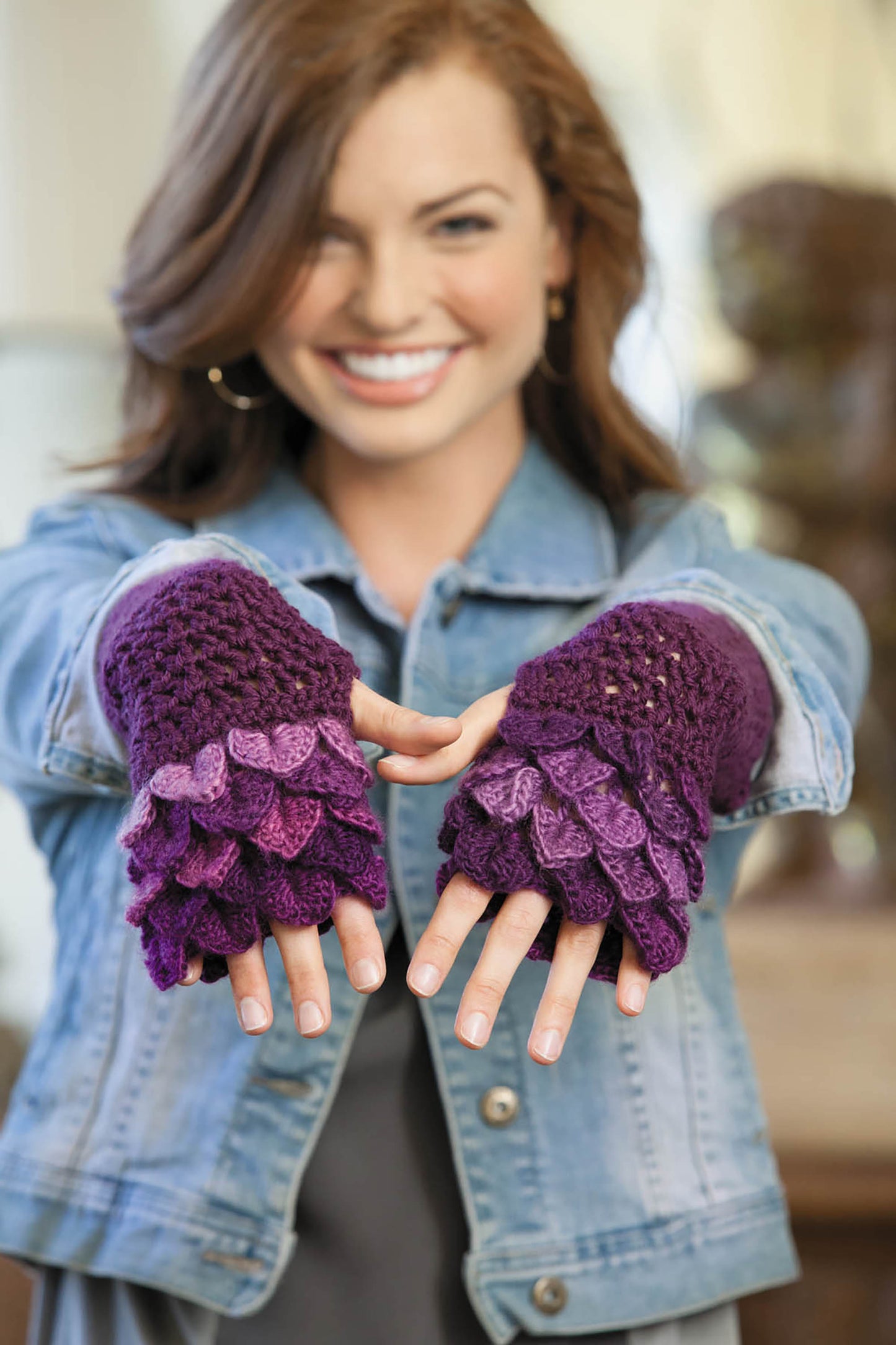 A smiling woman in a denim jacket shows off purple crochet fingerless gloves with scale-like patterns—made using the Crocodile Stitch from Leisure Arts' "Beginner's Guide to Crocodile Stitch," perfect for anyone seeking unique crochet patterns.