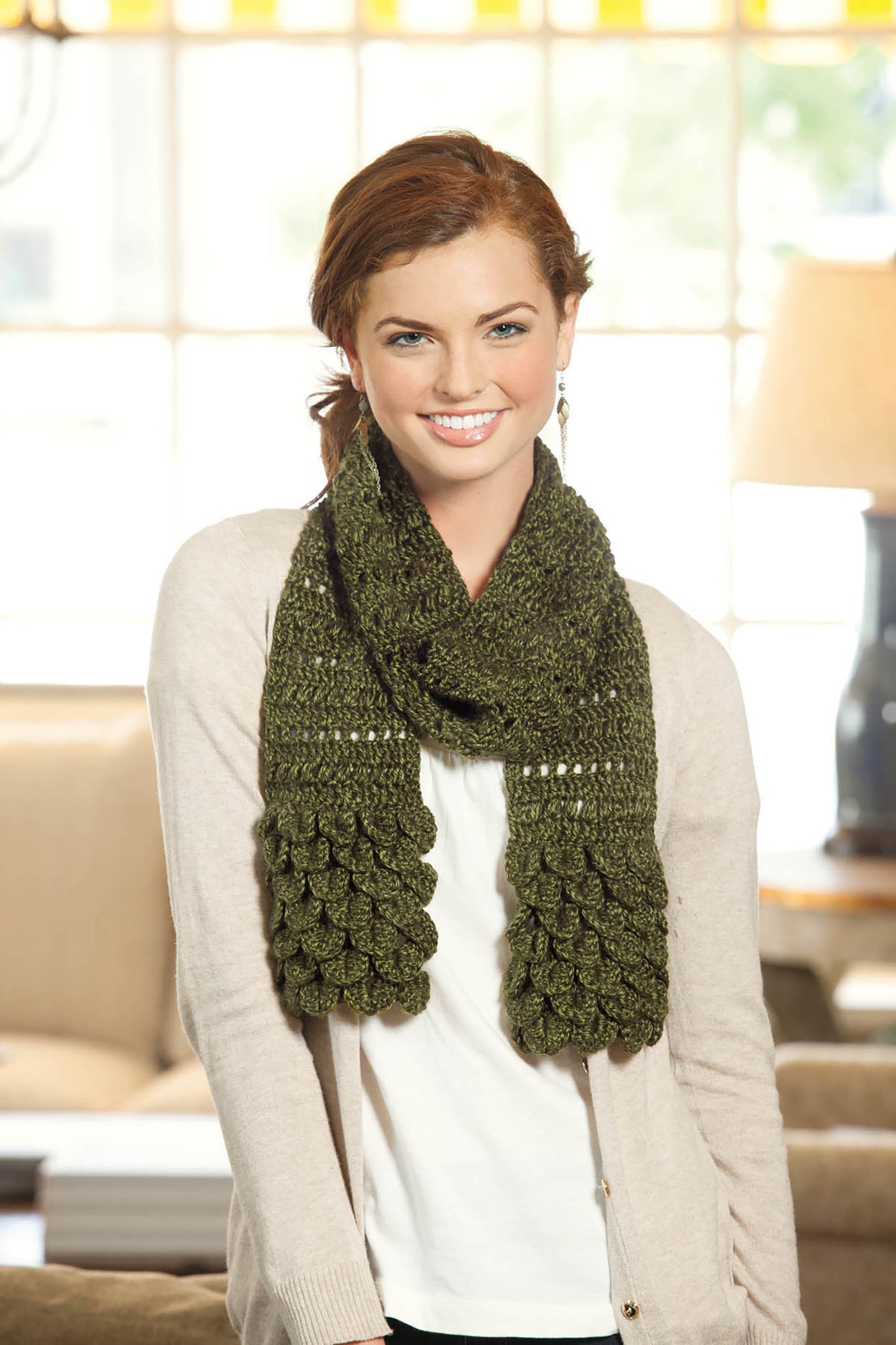 A red-haired woman smiles indoors, wearing a white top, beige cardigan, and a green scarf made using Crocodile Stitch from Leisure Arts' Beginner's Guide to Crocodile Stitch. She stands in a bright living room with large windows.
