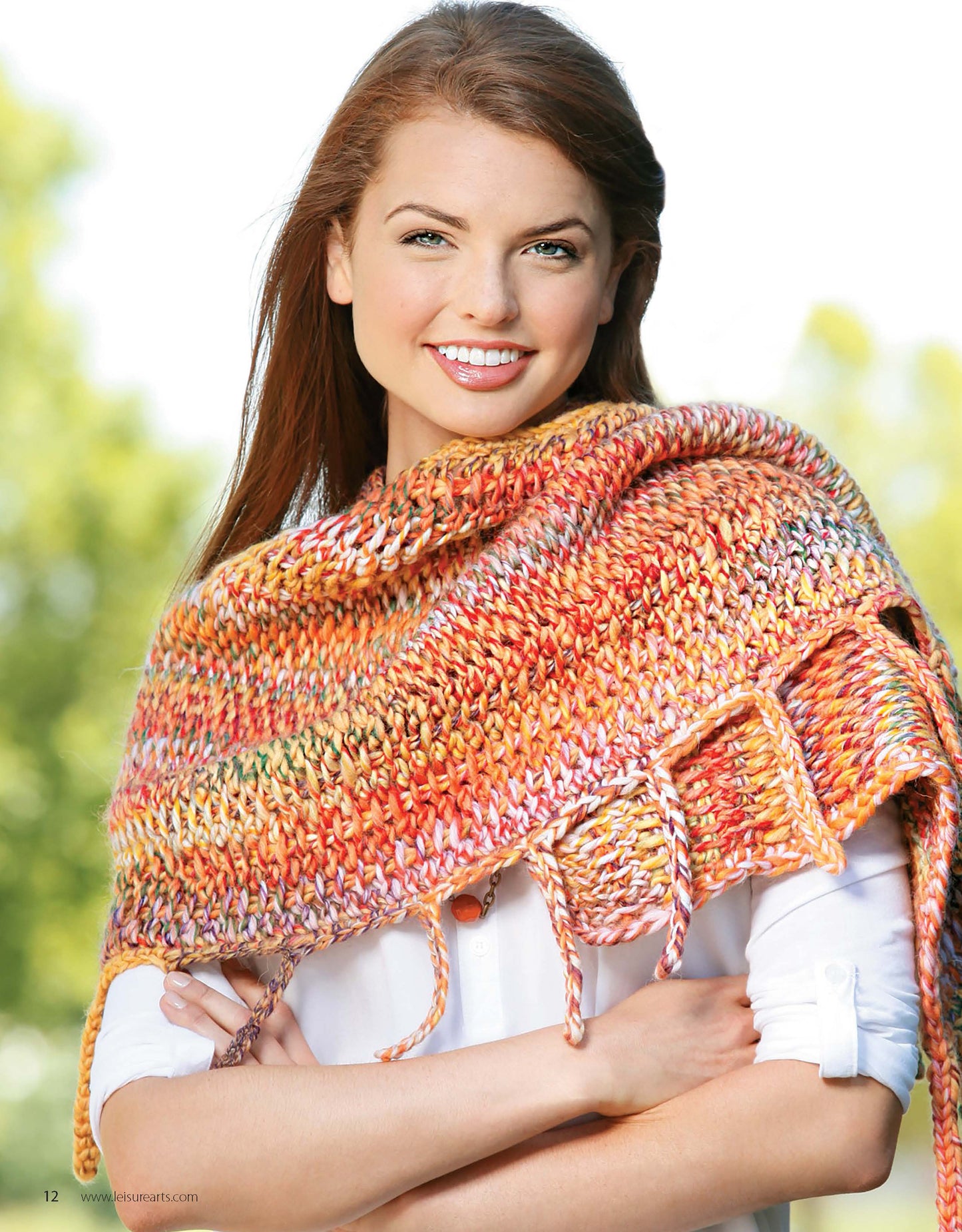 A woman with long brown hair smiles while modeling a Crochet Tunisian Shawl by Leisure Arts in orange and yellow over a white shirt, standing outdoors with arms gently crossed against a blurred green background.