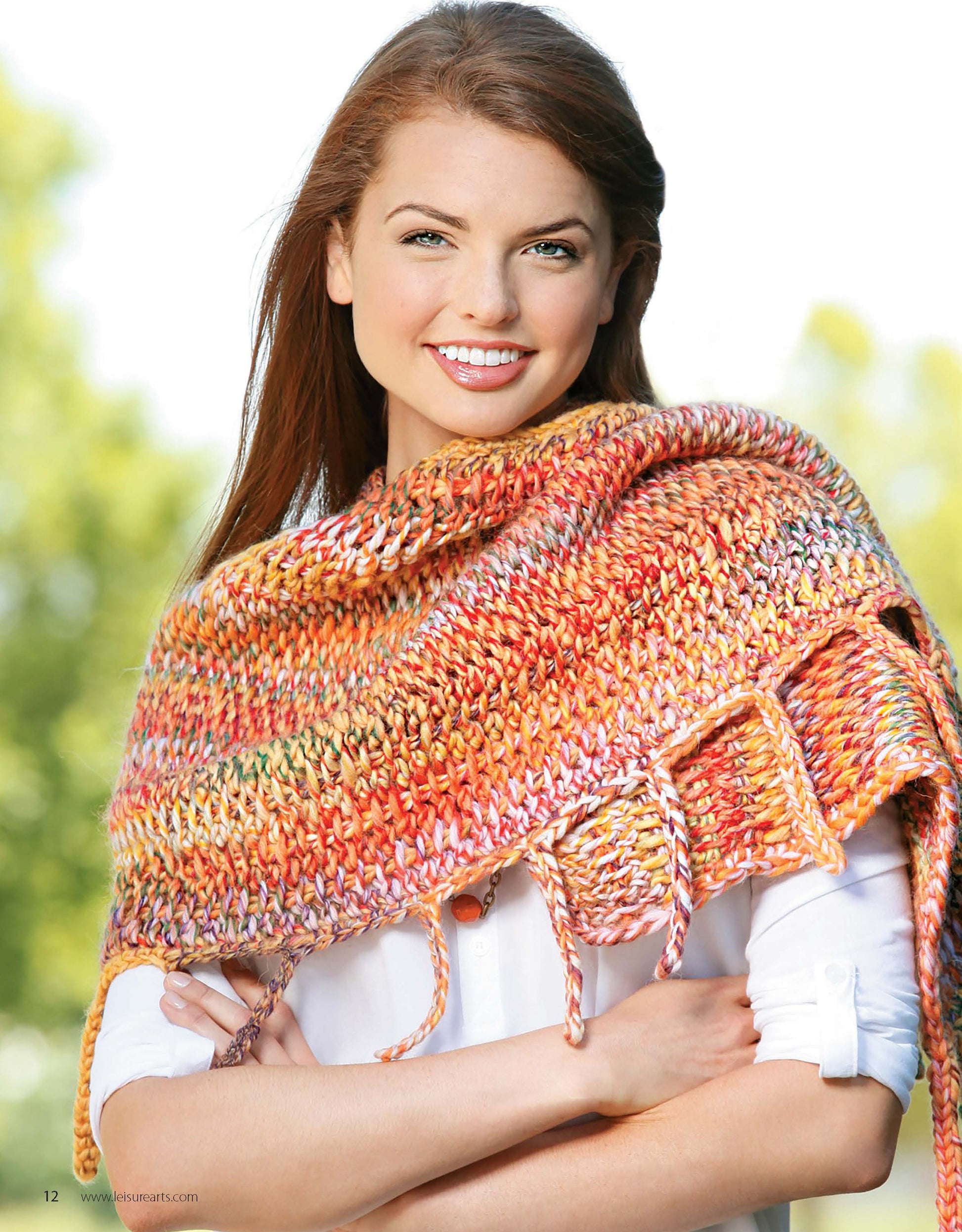 A woman with long brown hair smiles while modeling a Crochet Tunisian Shawl by Leisure Arts in orange and yellow over a white shirt, standing outdoors with arms gently crossed against a blurred green background.
