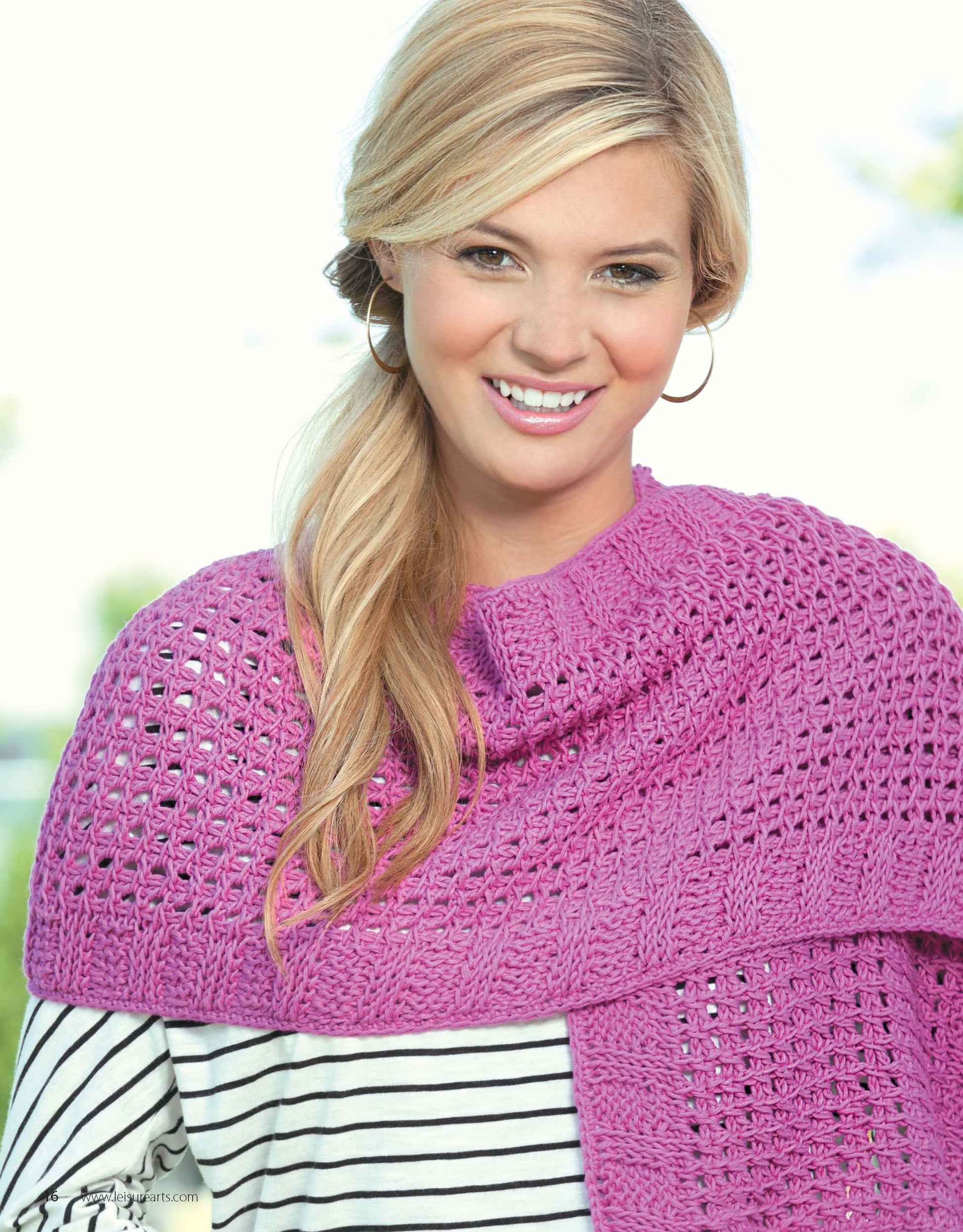 A smiling blonde woman wears large hoop earrings, a white and black striped shirt, and a textured magenta Crochet Tunisian Shawl by Leisure Arts draped over her shoulders. The softly blurred background shows greenery and sky.