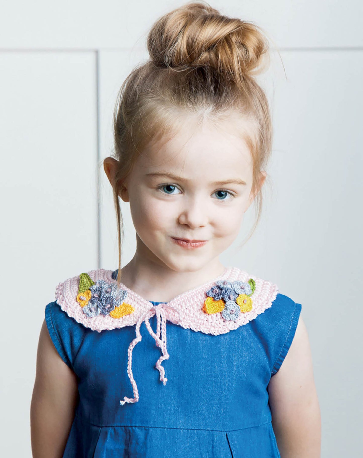 A young girl with light brown hair in a top bun models the Crochet Collars for Kids by Leisure Arts, featuring a blue dress accented with a pink crochet collar. She smiles gently and stands against a plain, light background.