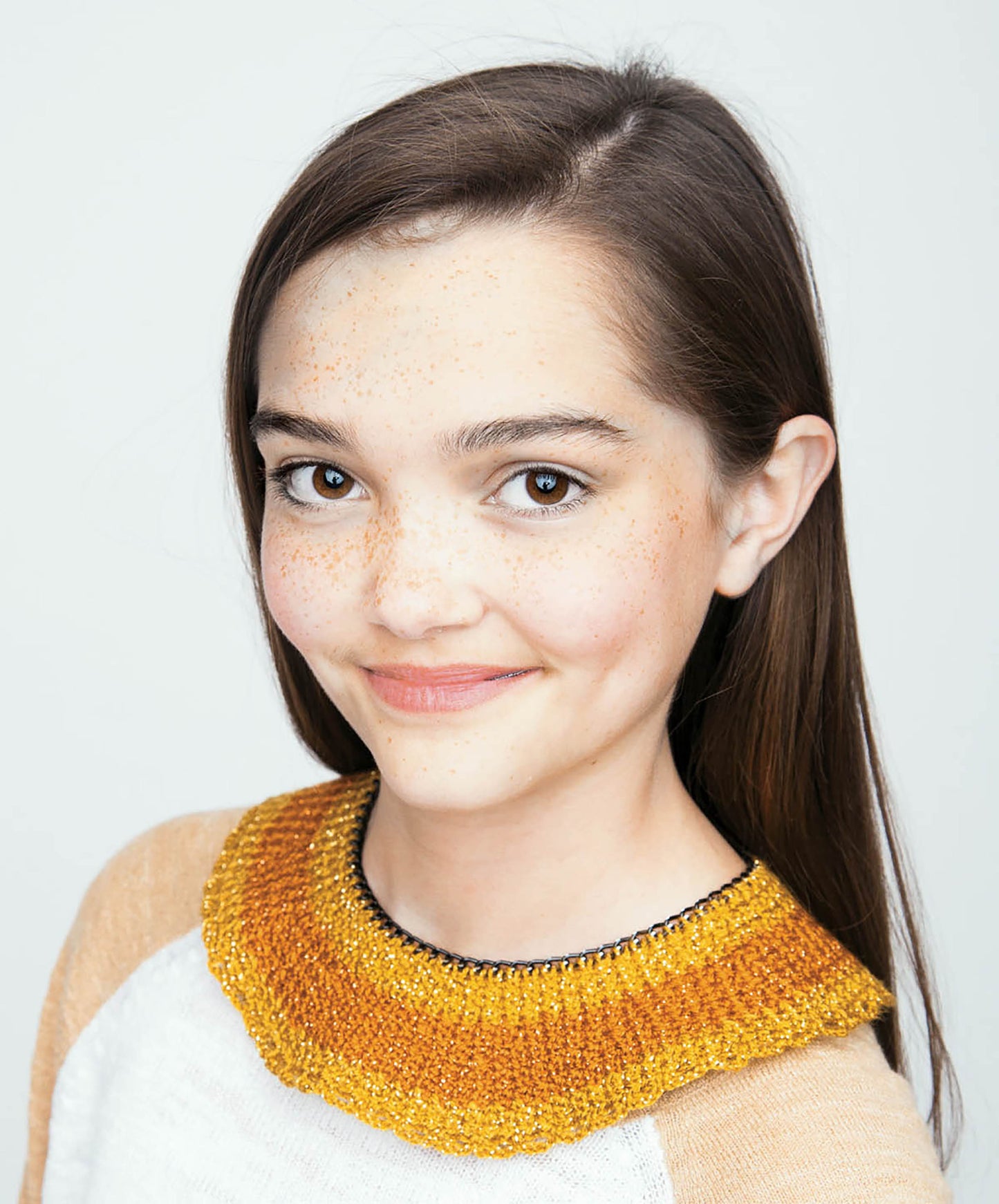 A young woman with straight brown hair and freckles smiles softly, modeling a chunky golden yellow crochet collar from Crochet Collars for Kids by Leisure Arts. She pairs it with a white top and tan sleeves against a plain, light background.
